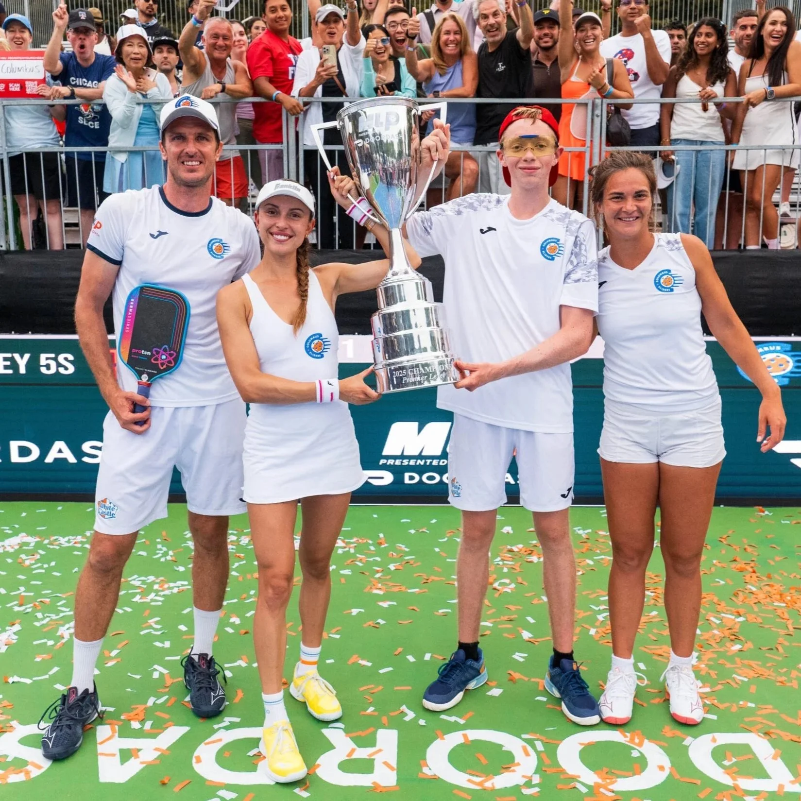 A group of four tennis players celebrating victory with a trophy on a tennis court, surrounded by a crowd of spectators.