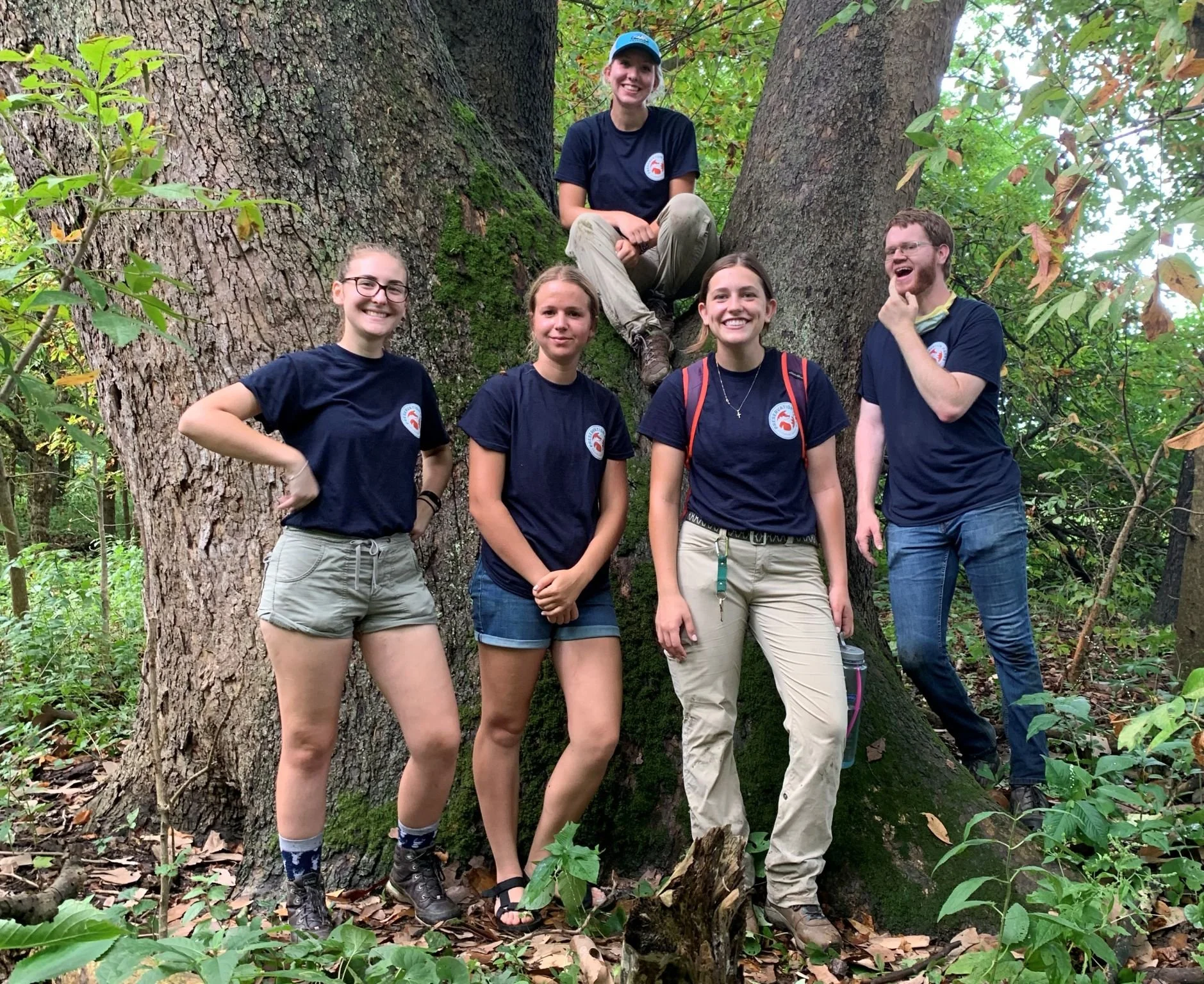 Group of five young people, four women and one man, standing and sitting around a large tree in a forest. They are all smiling and wearing matching navy T-shirts with a logo.