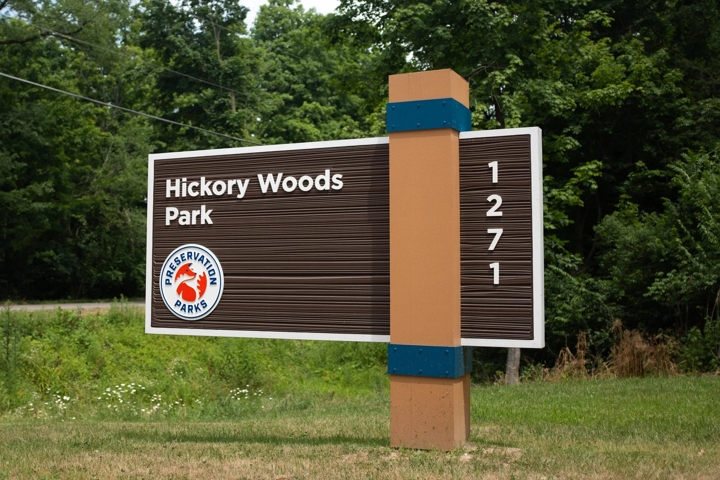 Sign for Hickory Woods Park with a reservation parks logo, located near a wooded area.
