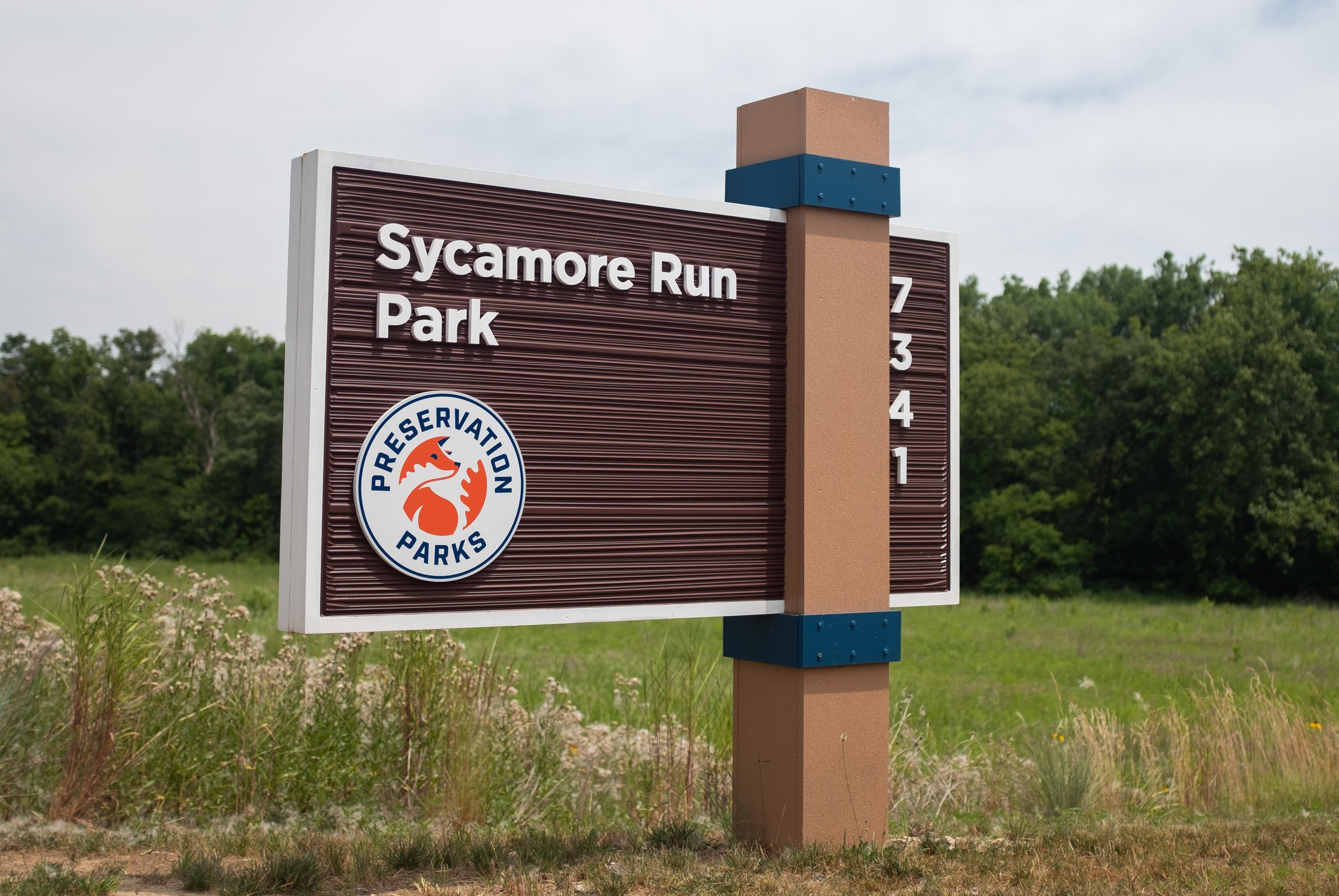 Sign for Sycamore Run Park, a Preservation Parks location, with address numbers 7, 3, 4, 1 visible, set in a grassy area with trees in the background.