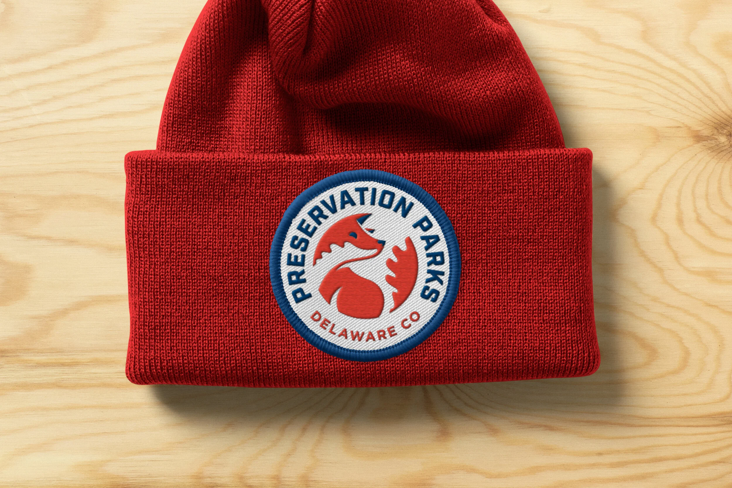 Red knit beanie hat with embroidered patch of the Preservation Parks Delaware Co logo on a light wood surface.