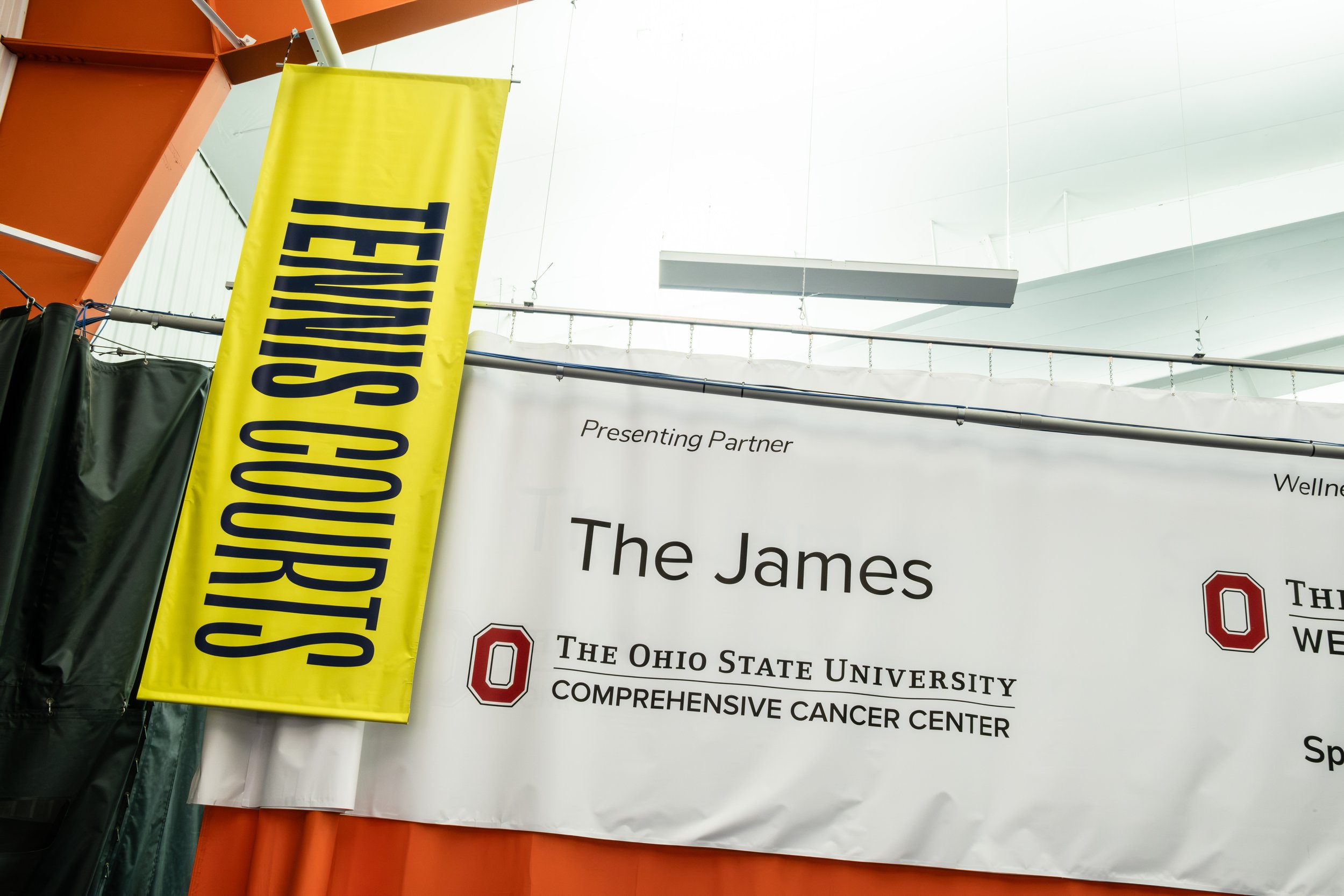 Exhibition banner promoting The Ohio State University Comprehensive Cancer Center, inside Pickle & Chill Indoor Pickleball with a smaller vertical yellow banner that says 'The Ohio State' in large black letters at a conference or event.