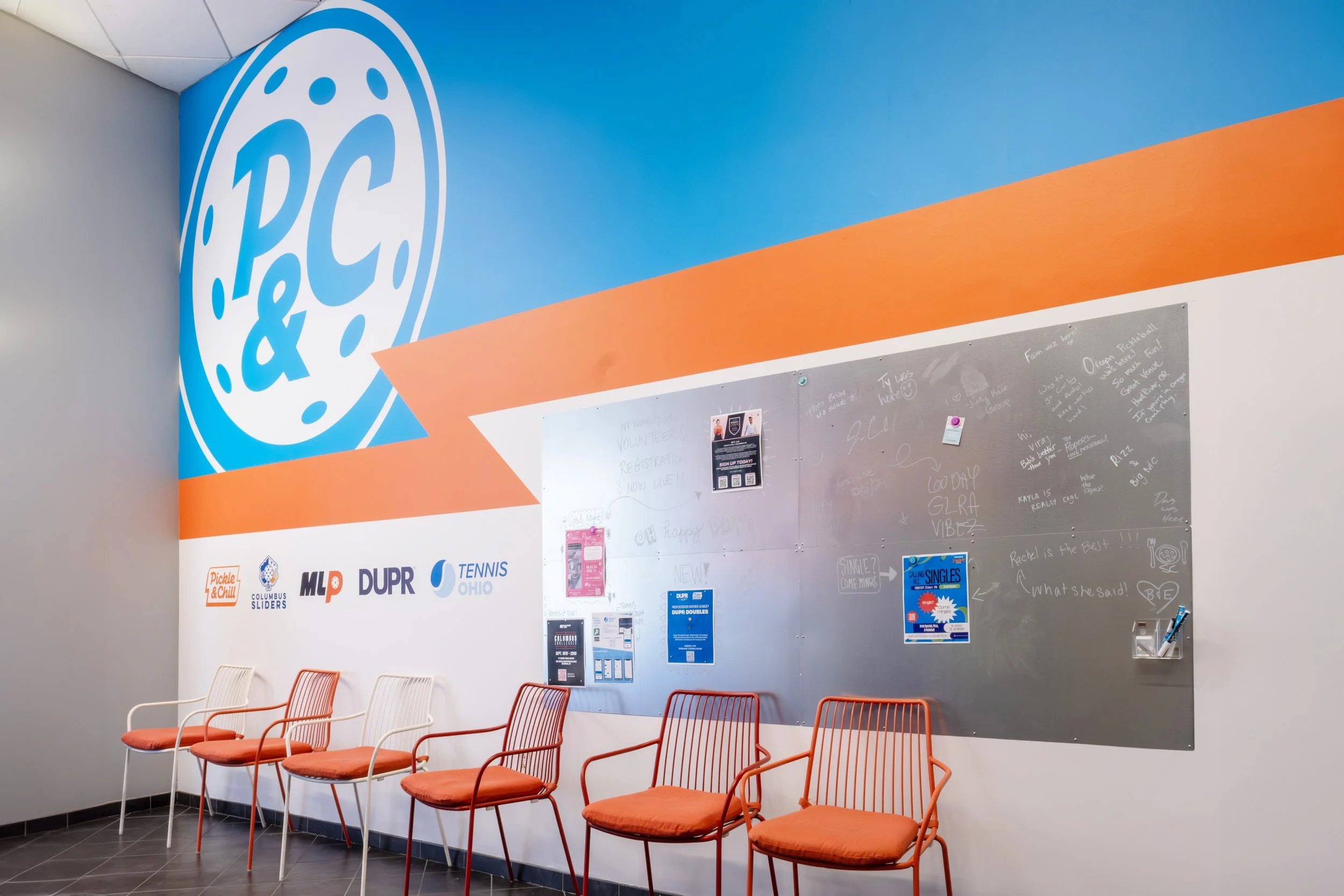 Indoor waiting area at Pickle & Chill Indoor Pickleball with five orange chairs against a wall. The wall features a large blue logo with the letters 'P&C' and various logos below it. There is a gray bulletin board with note paper and flyers pinned to