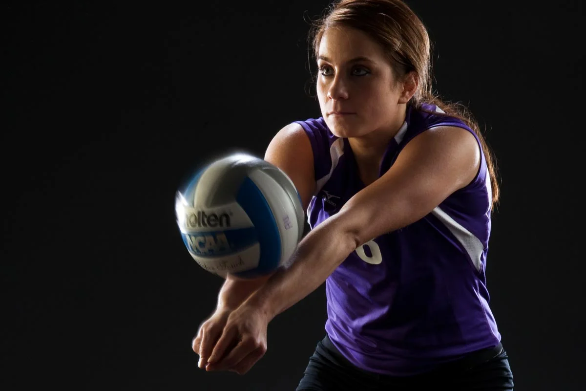 A female volleyball player in a purple uniform setting a volleyball against a dark background.