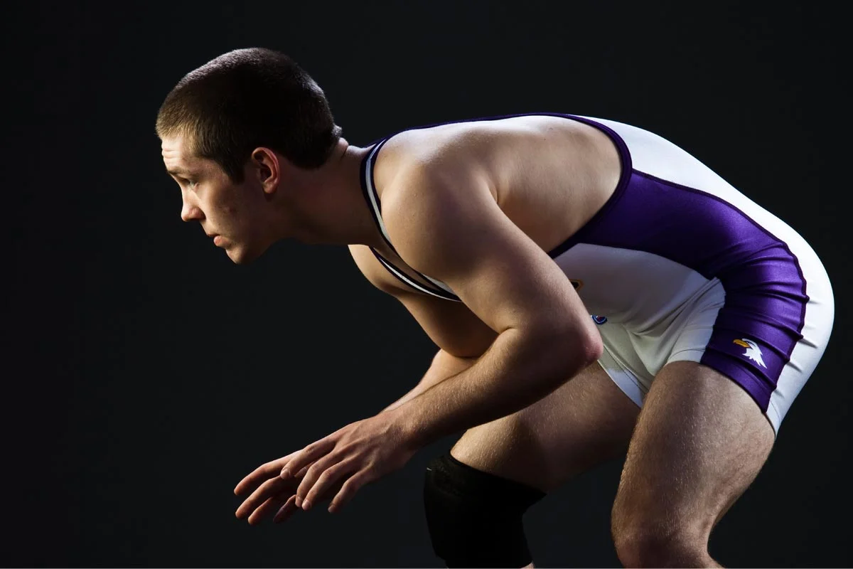 Young male athlete in a crouched position, wearing a purple and white sports uniform, against a dark background.