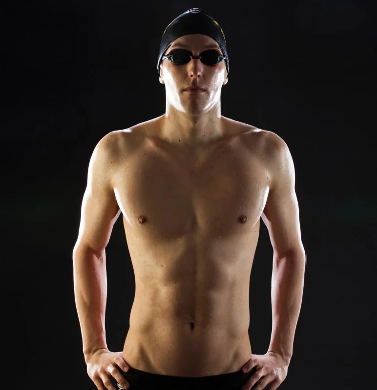 Male swimmer wearing a swim cap and goggles, shirtless, standing against a dark background.