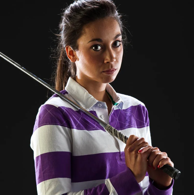 Young woman holding a golf club, dressed in a purple and white striped shirt, against a black background.