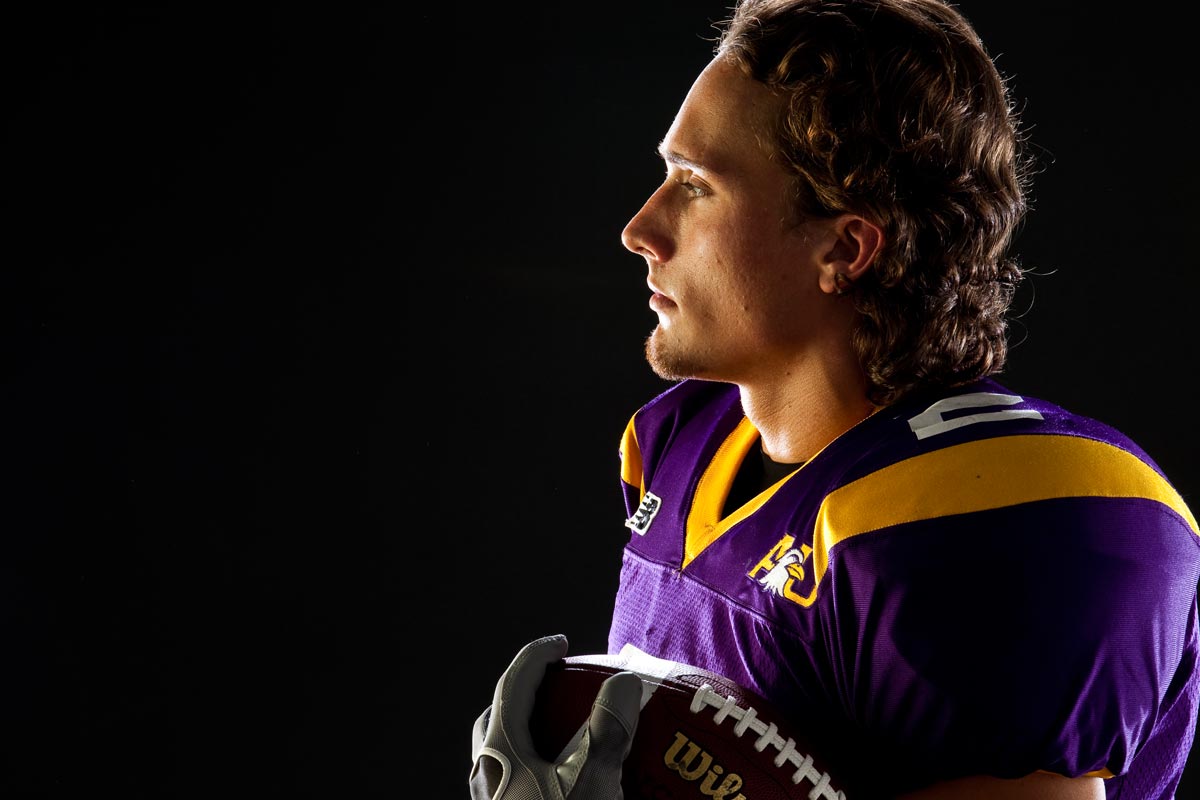 Side profile of a male football player in a purple and yellow uniform holding a football against a dark background.