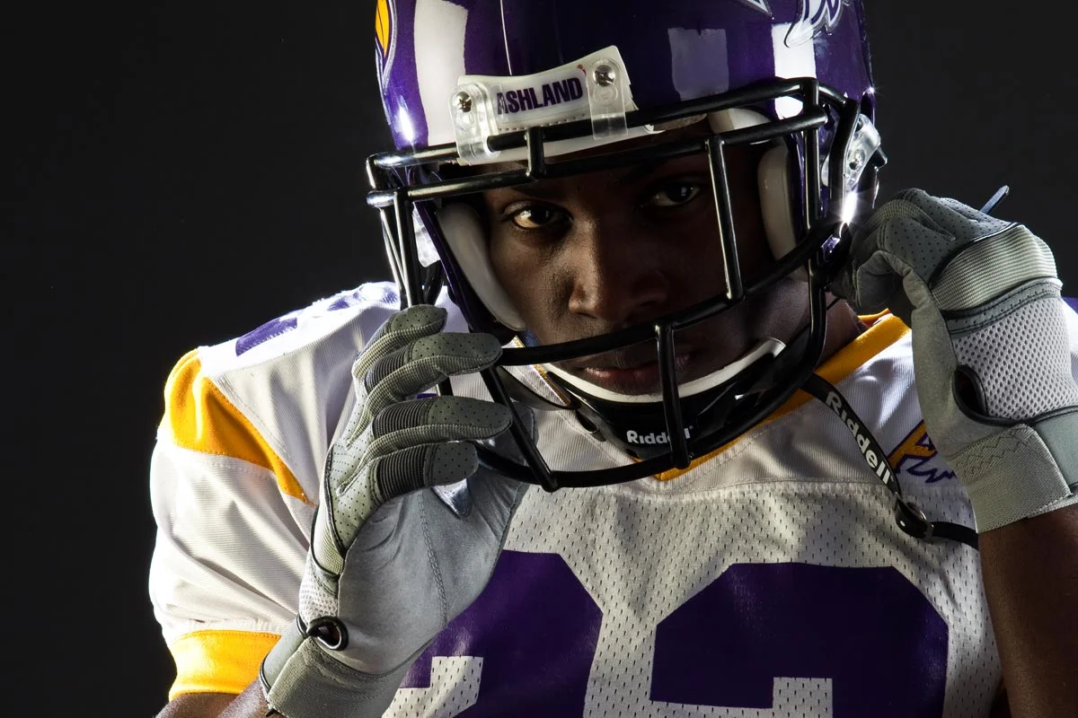 A football player wearing a purple and white helmet and jersey, adjusting his helmet. The player's intense gaze is visible through the face mask. The background is black.