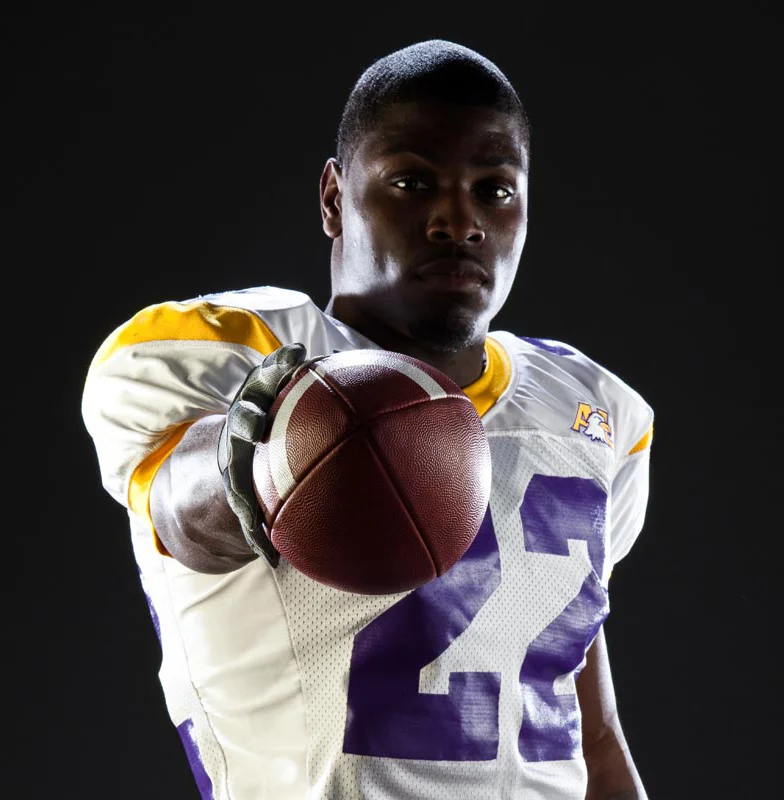 Football player in white and purple uniform holding a football extended toward the camera against a dark background.