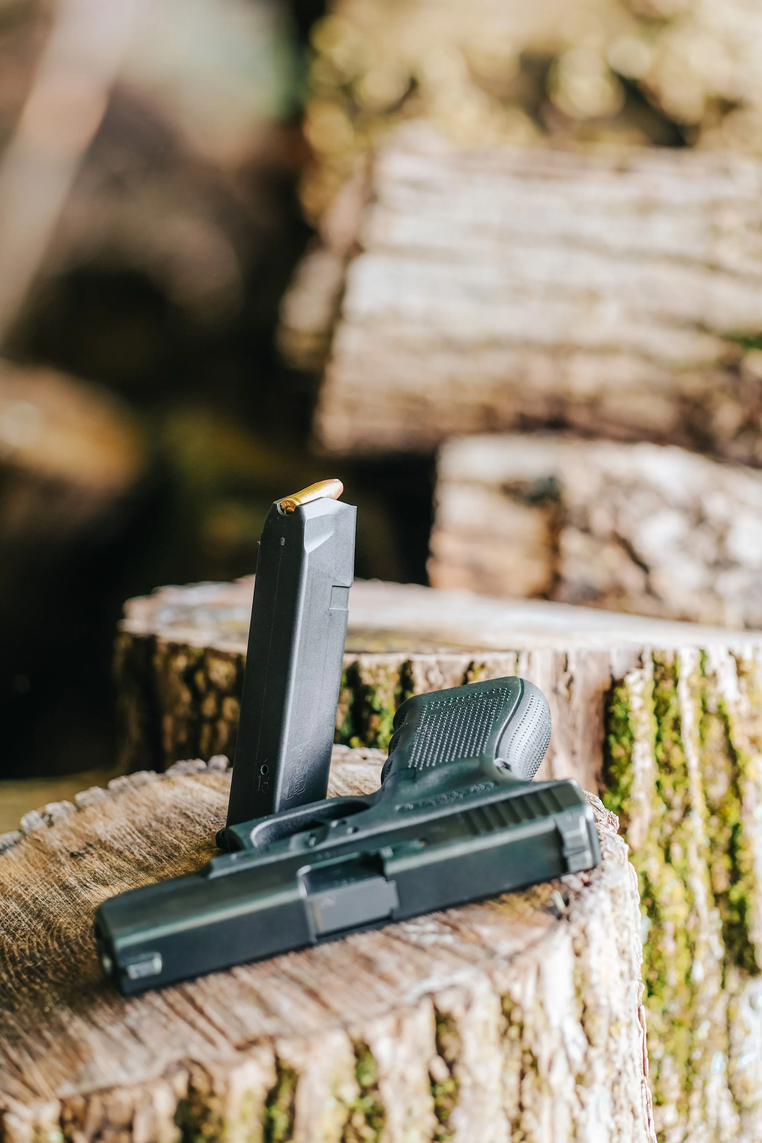 A handgun with a magazine inserted, lying on a tree stump with a bullet resting on top of the magazine, outdoors with blurred logs in the background.