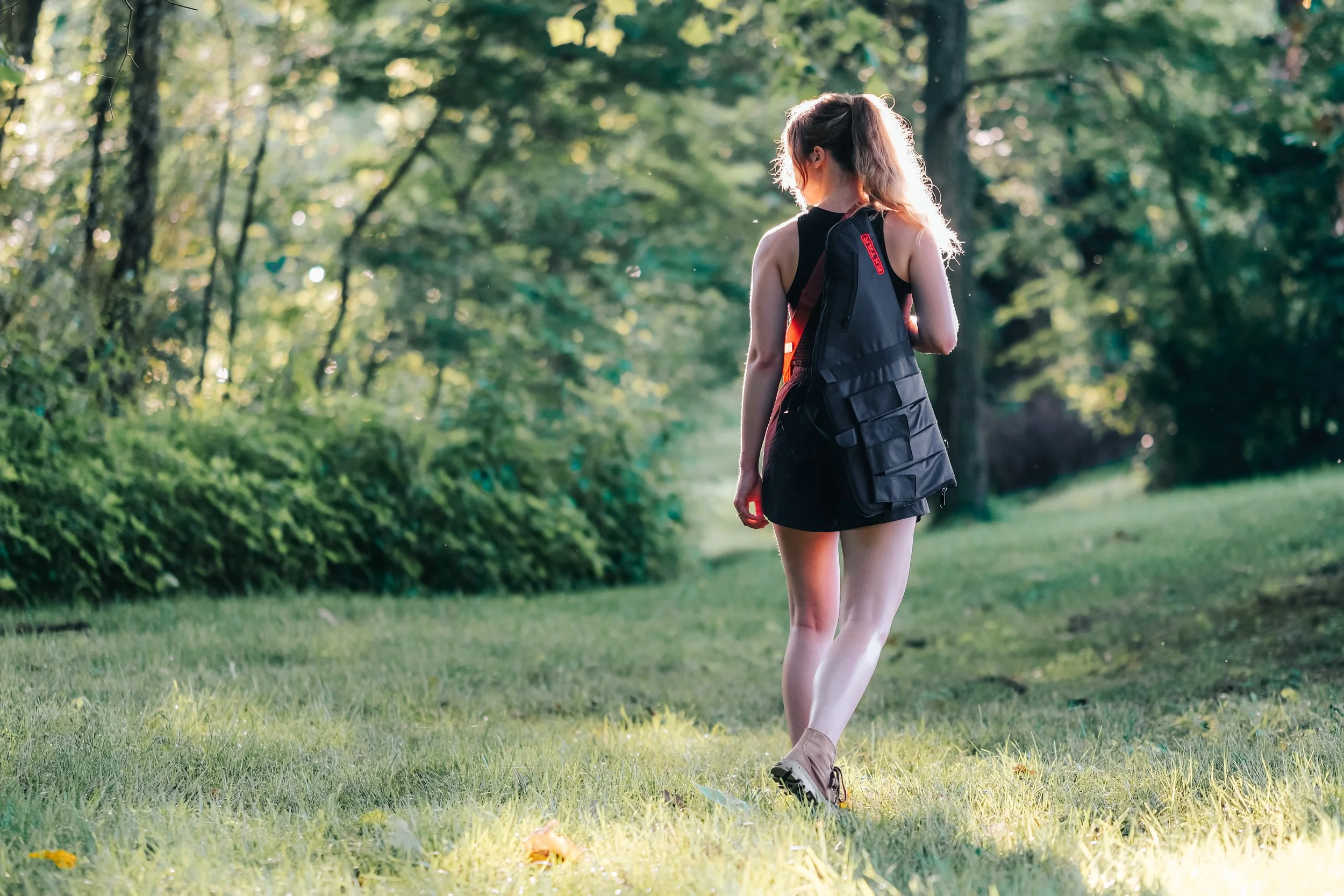 Young woman with long hair walking in a park during daytime, carrying a black backpack, wearing a black dress and hiking shoes.