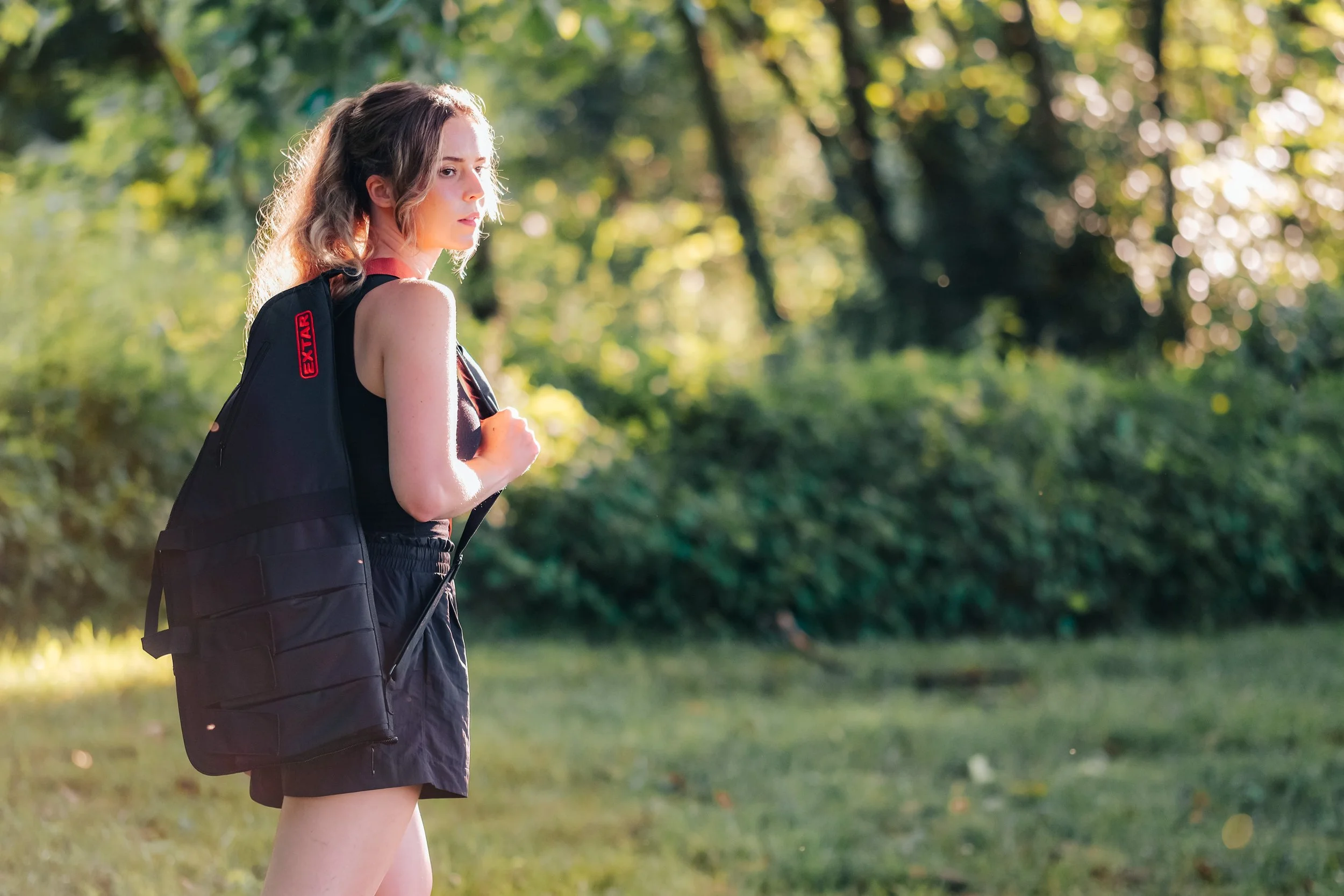 Young woman with long wavy hair carrying a black backpack outdoors in a green park during daytime.