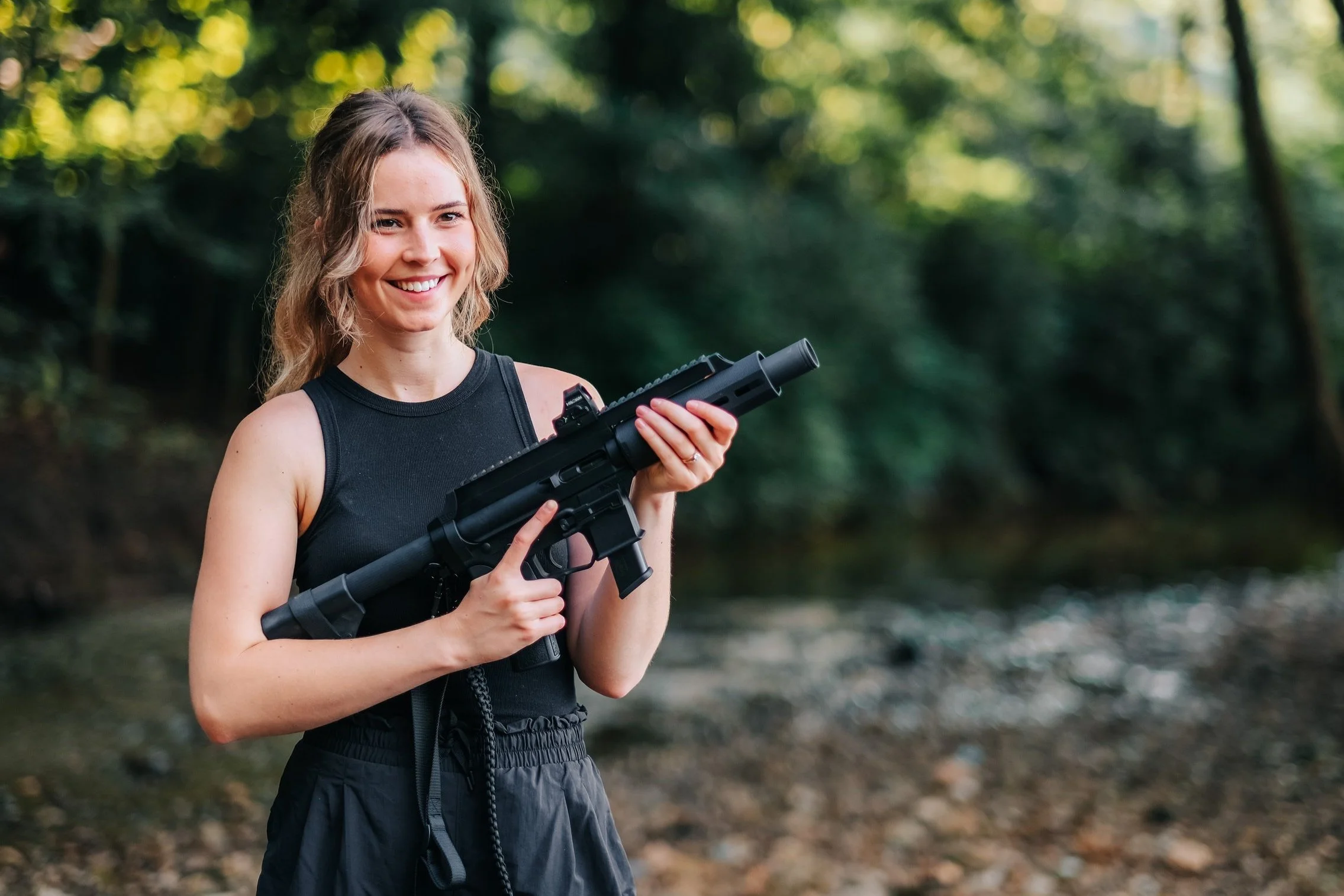 A smiling young woman with wavy blonde hair, wearing a black sleeveless top and black pants, outdoors in a wooded area, holding a black semi-automatic rifle.