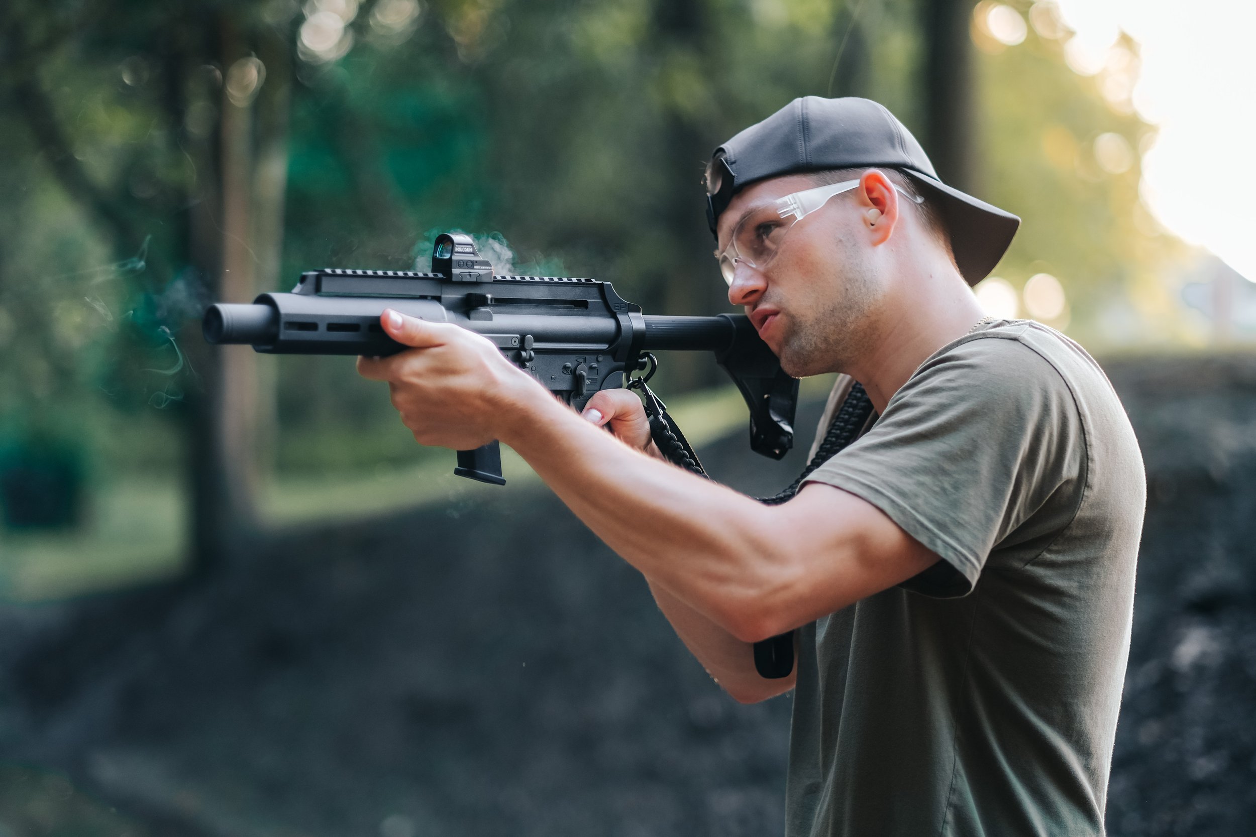 A man wearing a gray cap, safety glasses, and a khaki T-shirt aims and shoots a black rifle outdoors with trees in the background.