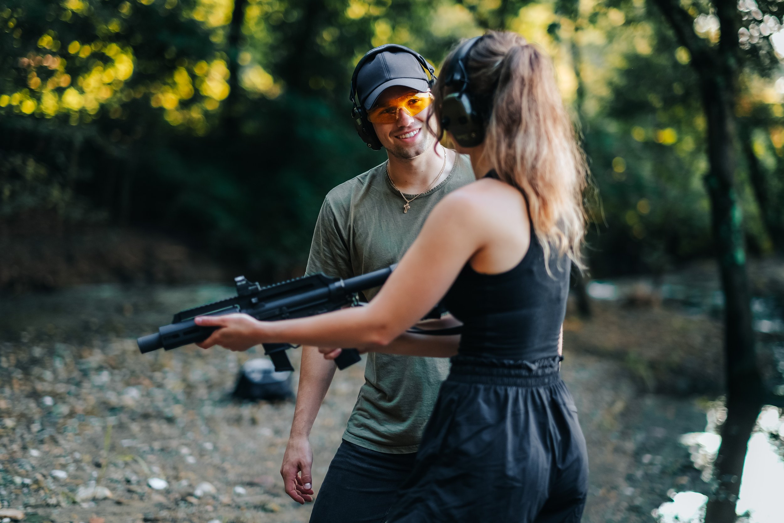 A man and a woman outdoors in a wooded area, the woman is holding a rifle, and the man is smiling and wearing safety glasses and headphones.