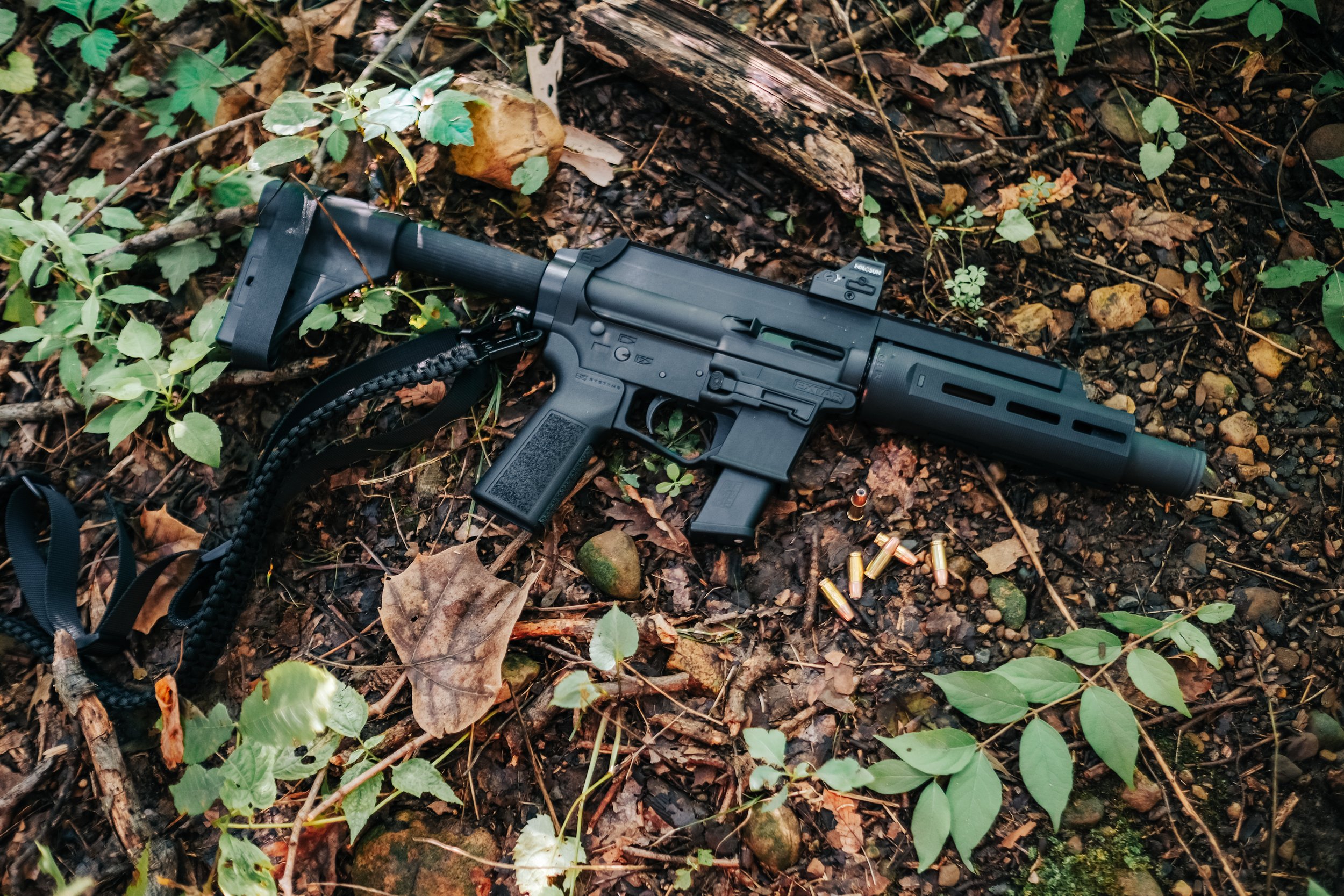 A black firearm with a suppressor, lying on a forest ground among leaves, twigs, and some spent ammunition shells.