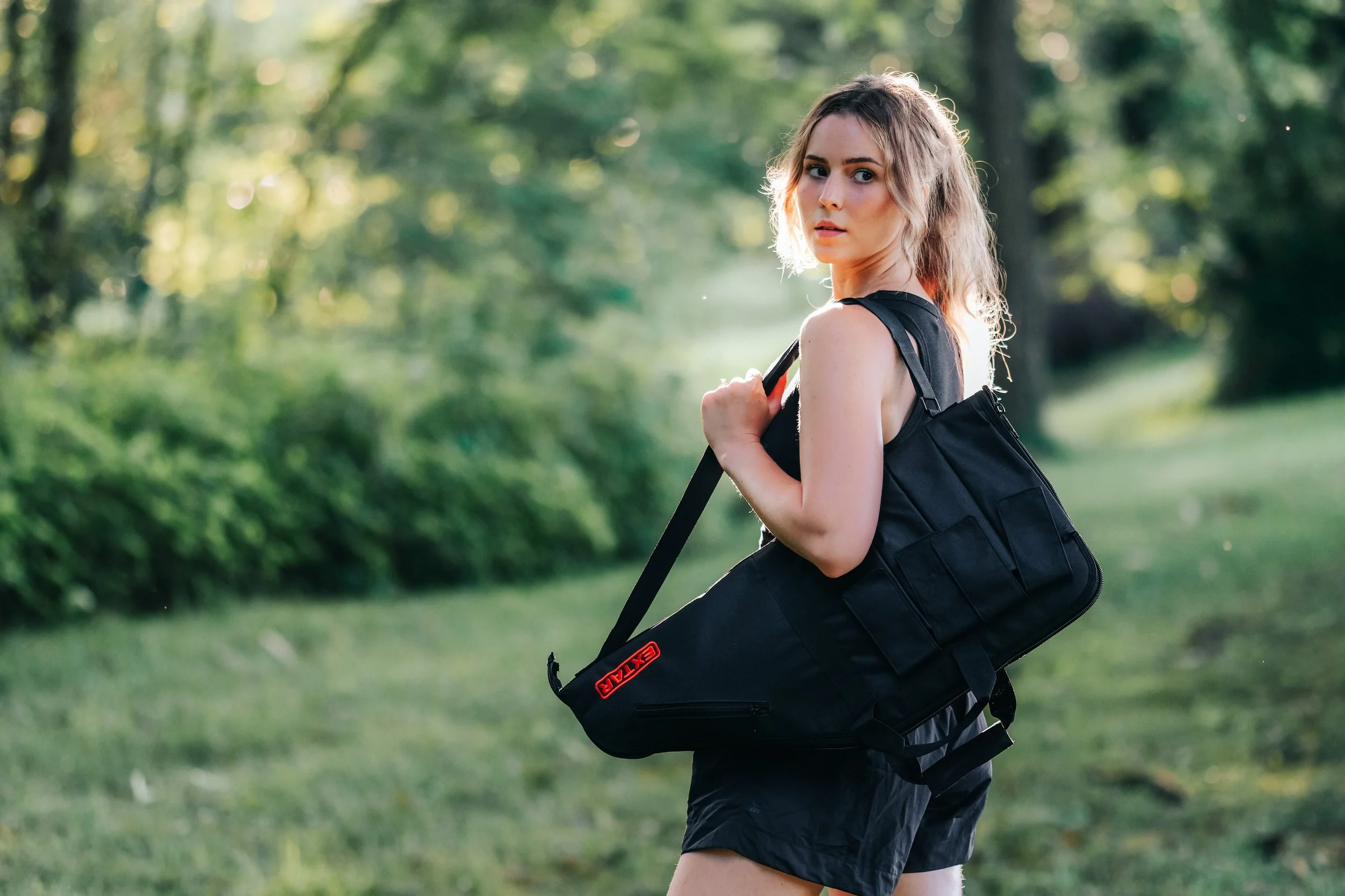 Young woman with wavy hair carrying a large black bag outdoors in a green park.