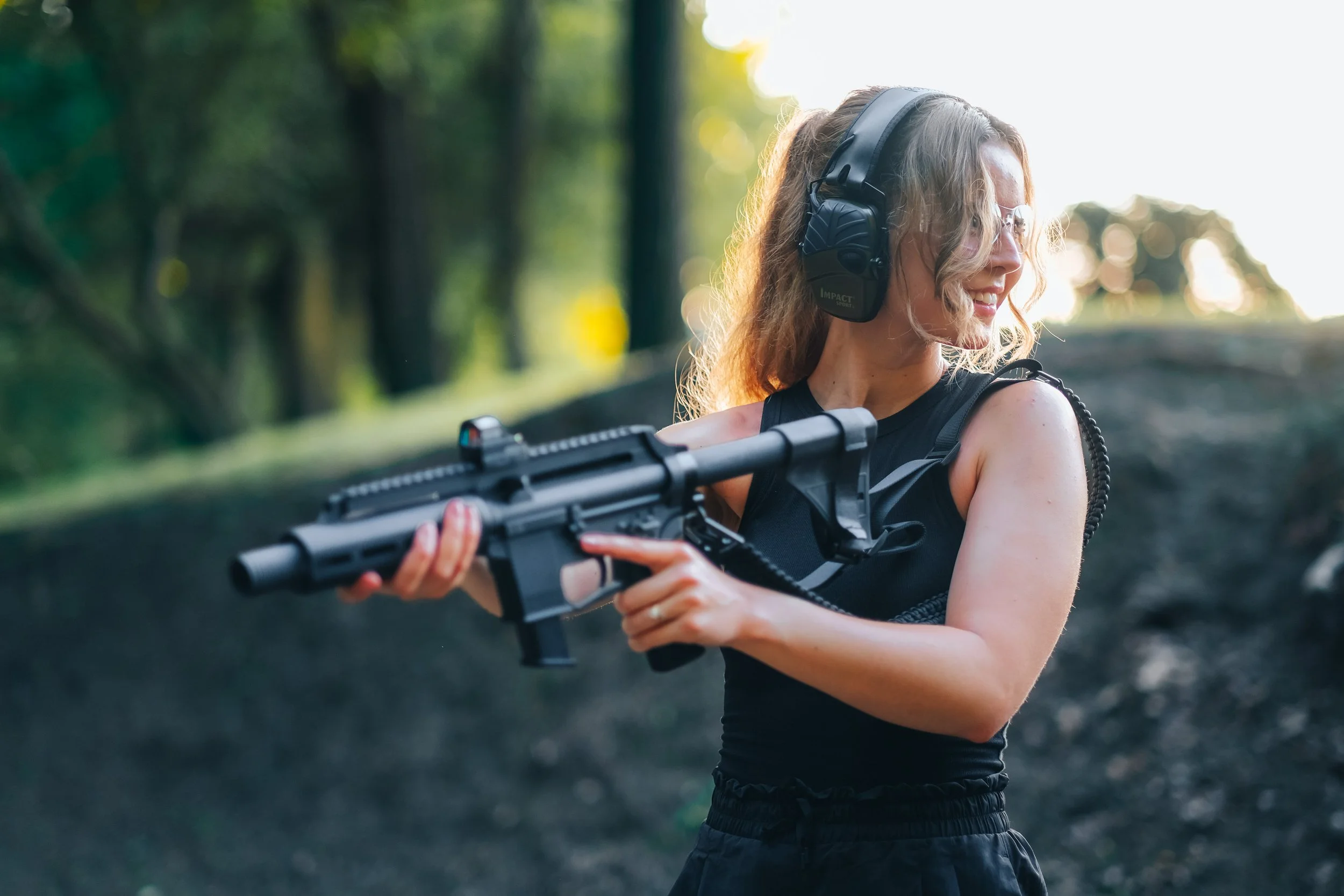 A woman outdoors holding a firearm, wearing headphones and protective eyewear, with trees and sunlight in the background.
