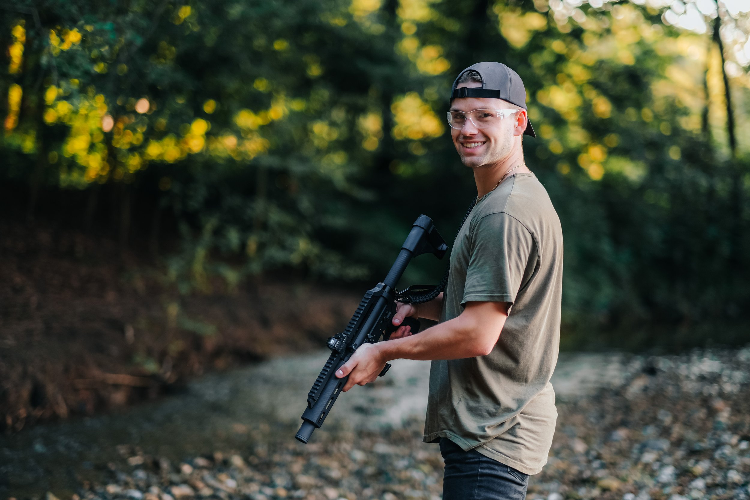 A young man wearing a backward cap, glasses, and a green t-shirt standing outdoors near a creek, holding a large black rifle and smiling at the camera.