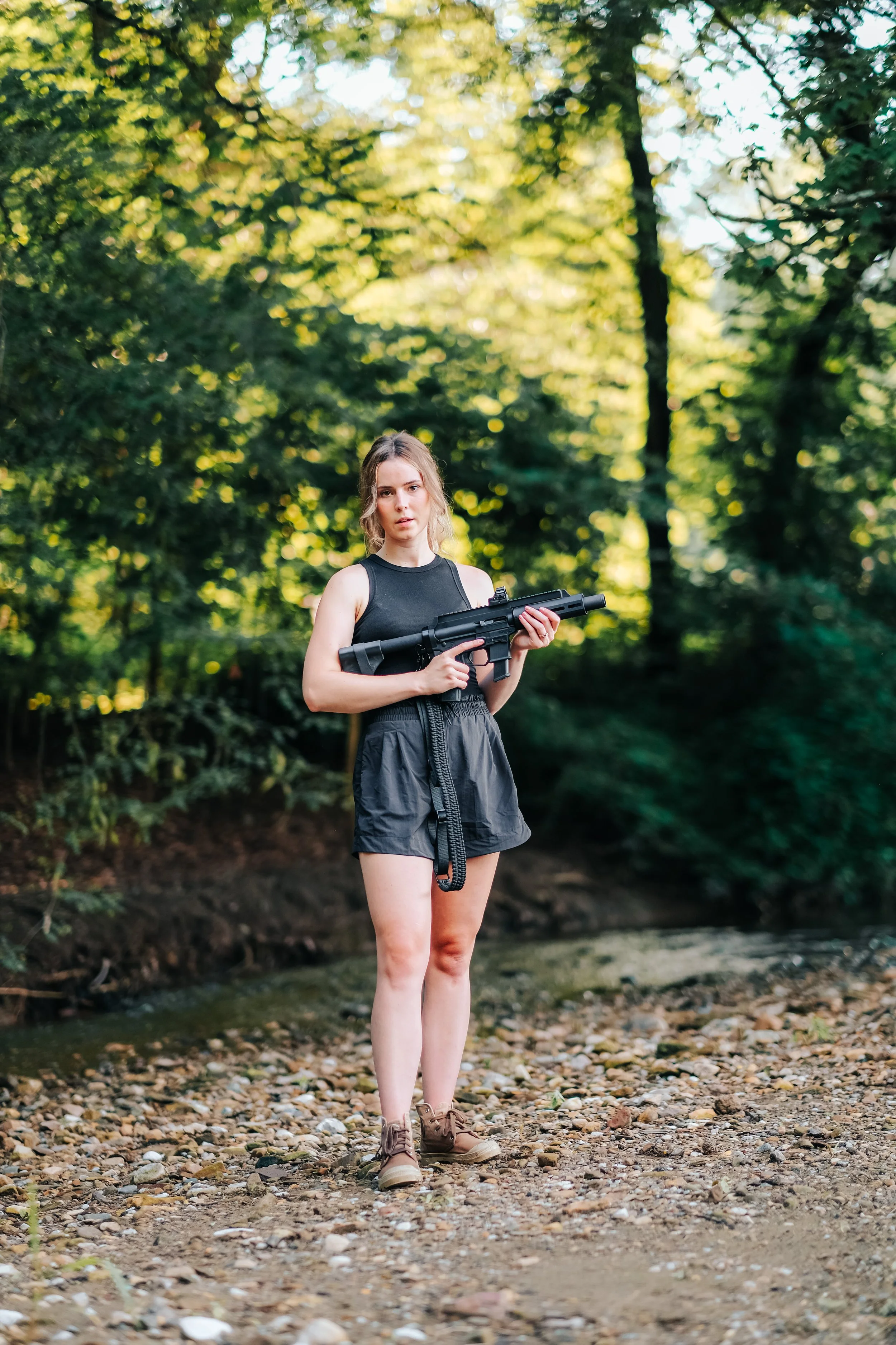 A young woman standing outdoors on a rocky path holding a large black gun, wearing a black sleeveless top, black shorts, and brown boots, with green trees and sunlight in the background.