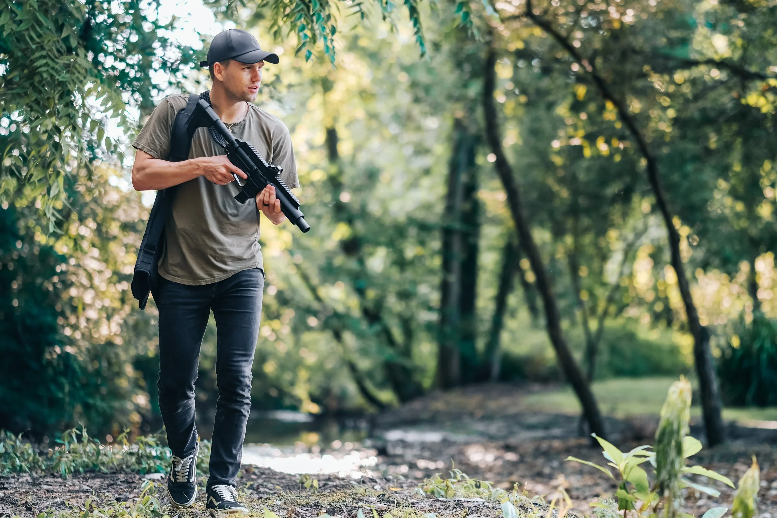 A man wearing a gray t-shirt, black jeans, and a black cap, walking through a dense forest with a rifle in his hands.