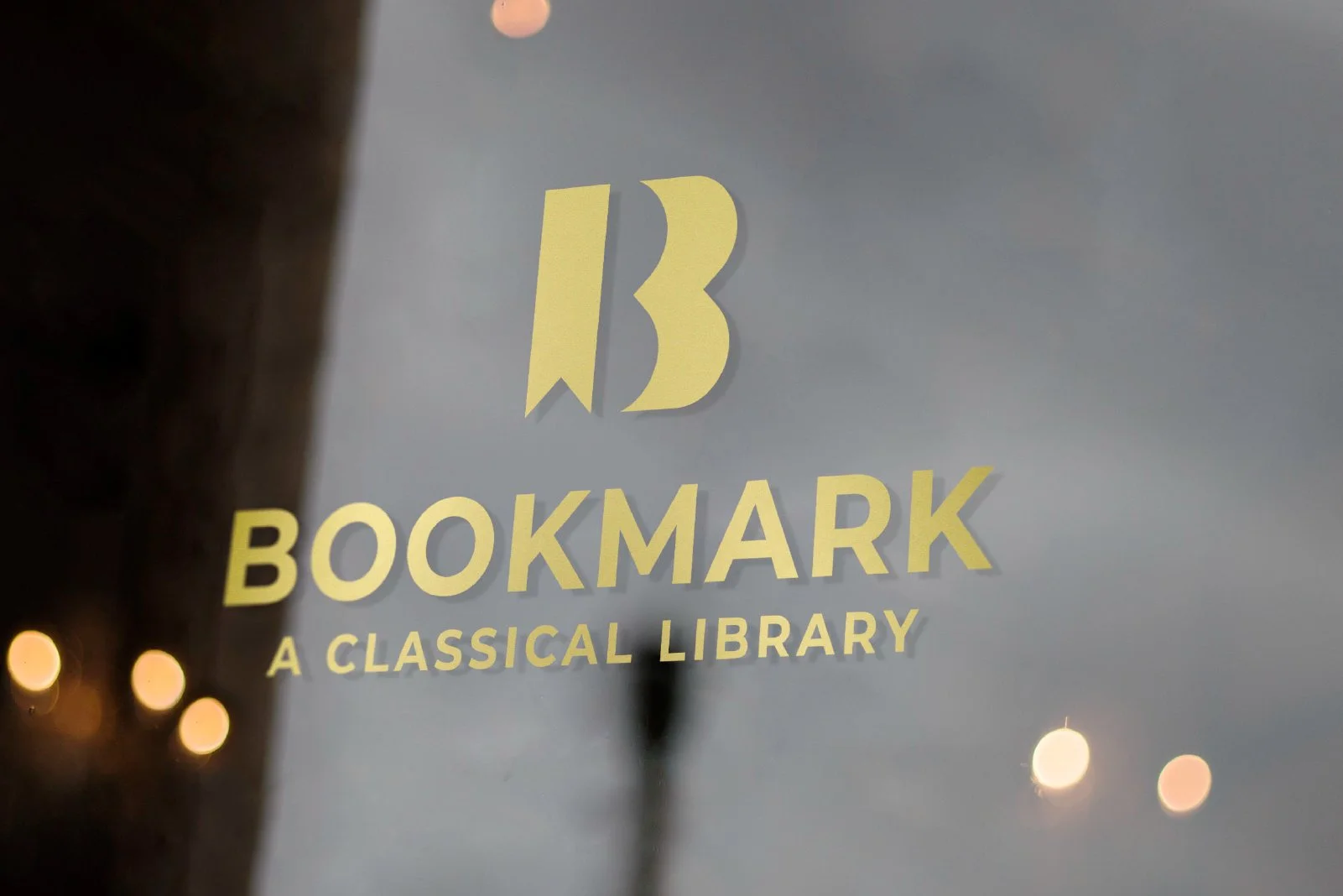 Sign for Bookmark, a classical library, with the logo and name in yellow lettering on a glass window, with blurry city lights reflected.
