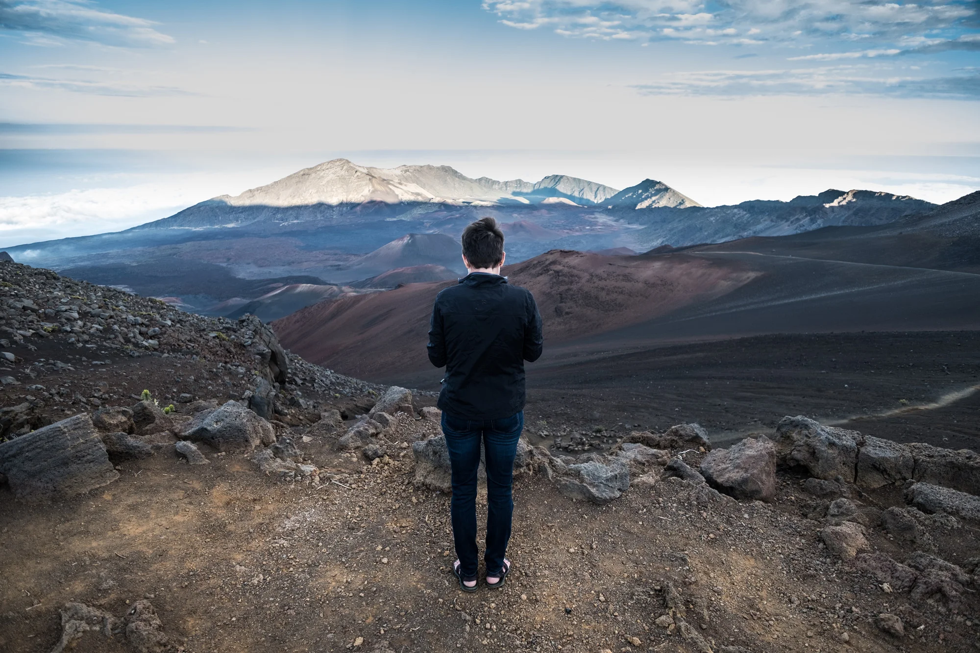 A person standing on rocky terrain, facing away, overlooking a mountainous landscape with volcanic mountains under a partly cloudy sky Maui.