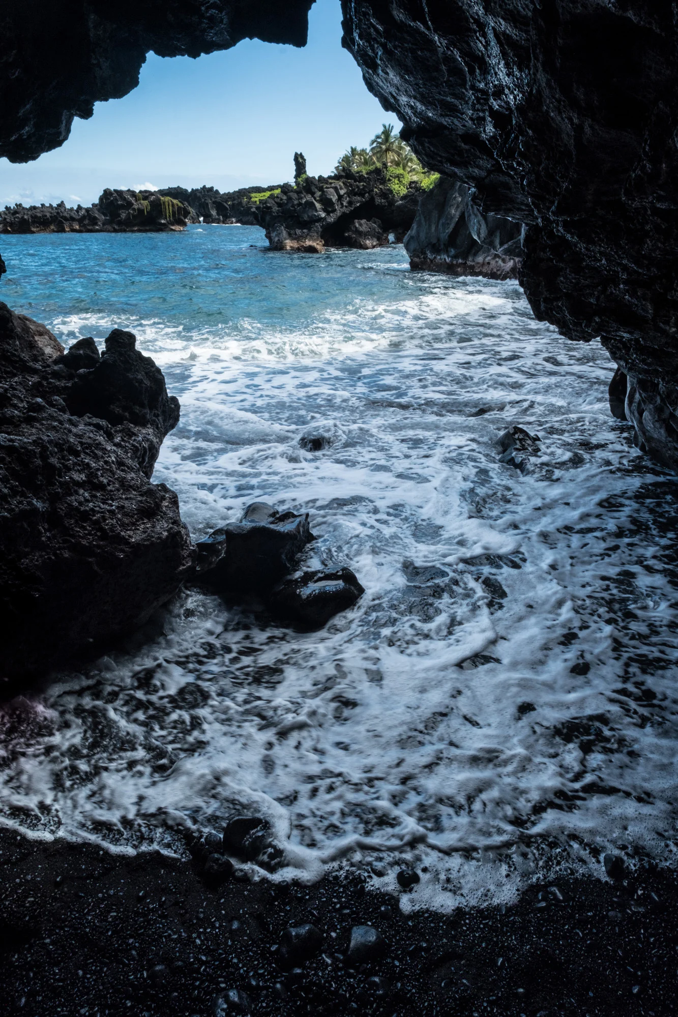 View from inside a sea cave looking out at the ocean with waves crashing near dark rocks. In the distance, there's a small island with palm trees and lush vegetation in Maui.