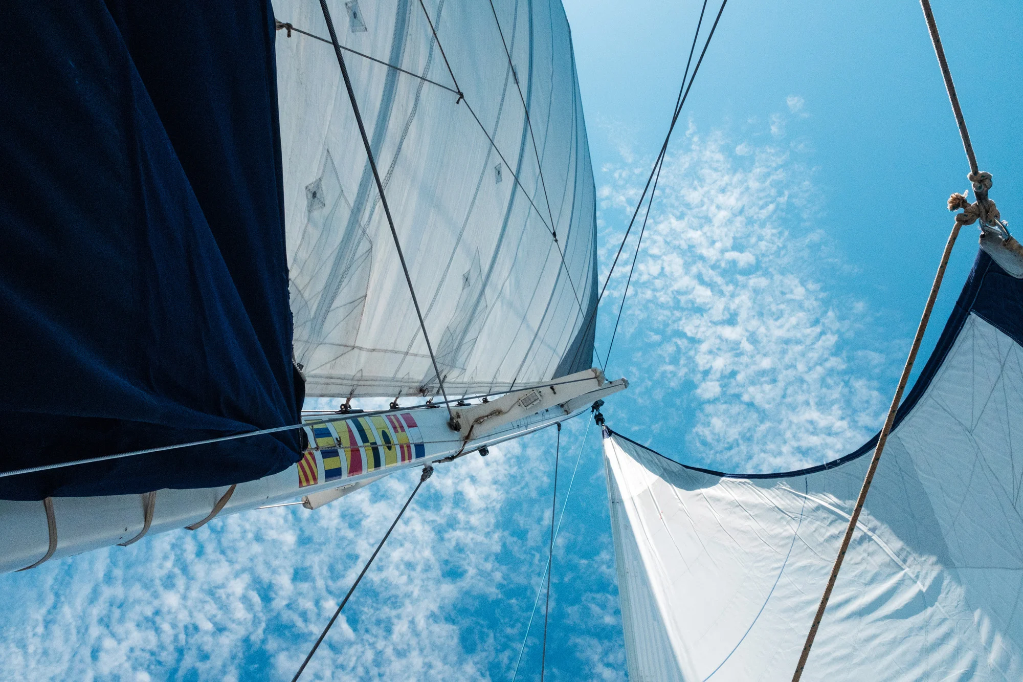 A view looking up at a sailboat's mast and sails with a bright blue sky and scattered clouds in the background in Maui.