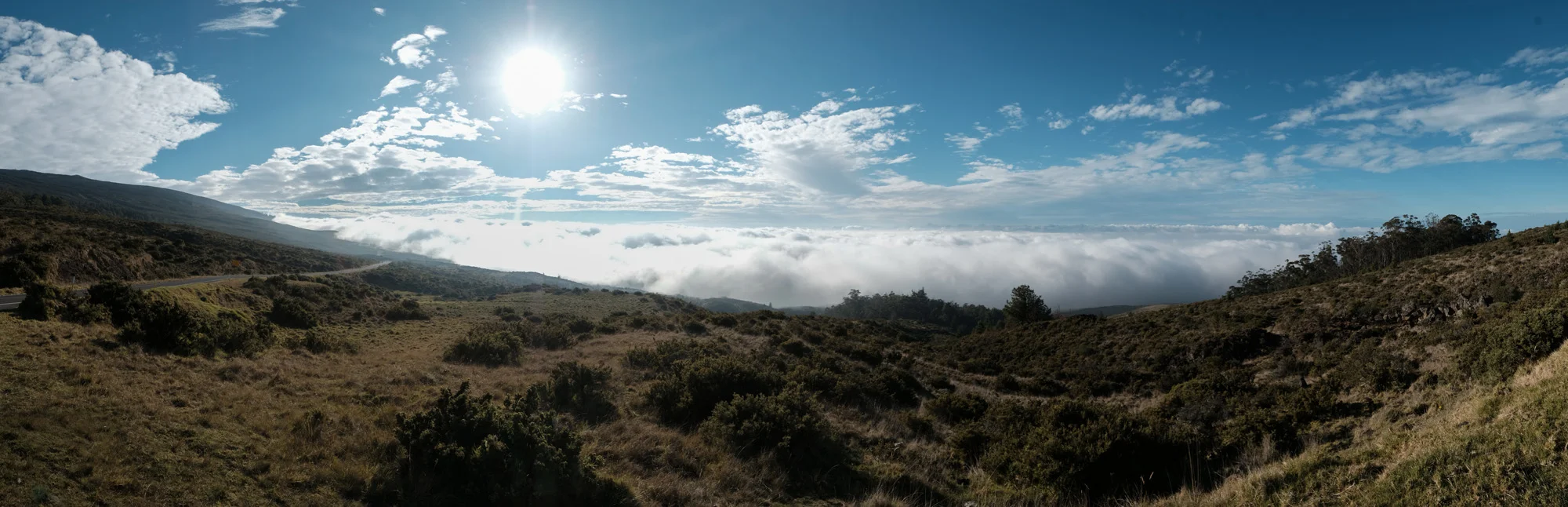 A scenic landscape of rolling hills with dry grass and sparse bushes, a road on the left, clouds below the horizon, and a bright sun shining in a partly cloudy sky Maui.