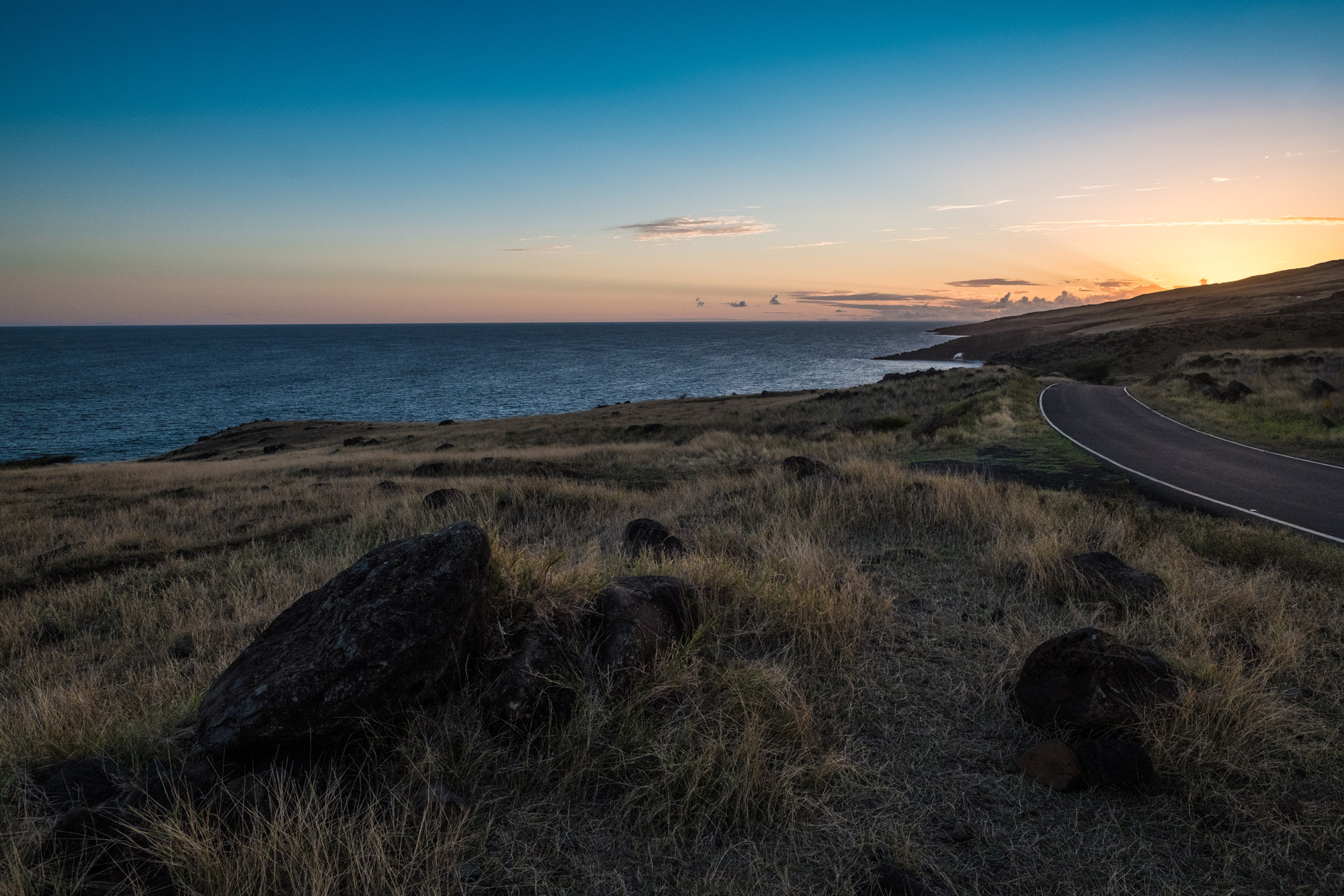 A coastal landscape at sunset, featuring a winding road along grassy terrain, cliffs, and the ocean on the horizon in Maui.