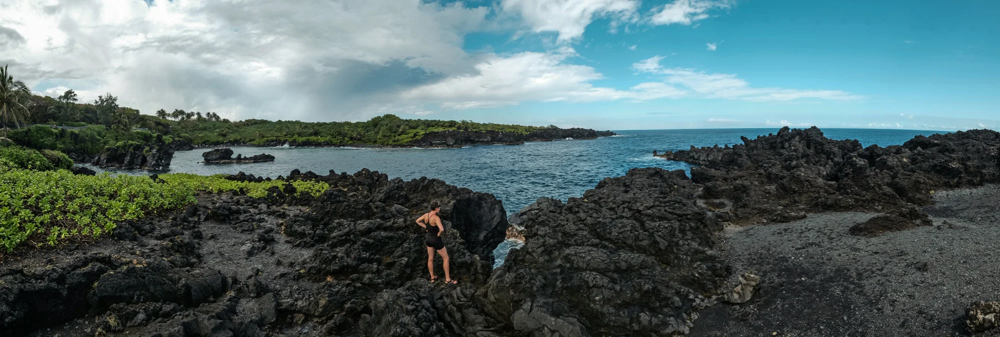 Woman standing on black volcanic rocks overlooking the ocean with cloudy sky and green trees in the background in Maui.