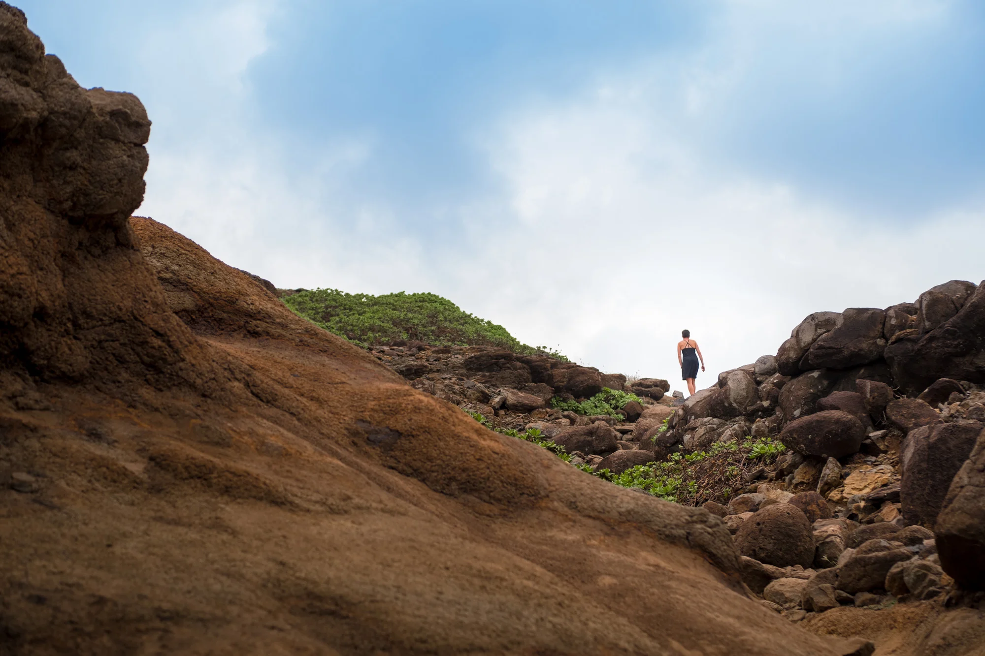 A person walking along a rocky coastline with large rocks and greenery under a cloudy sky Maui.