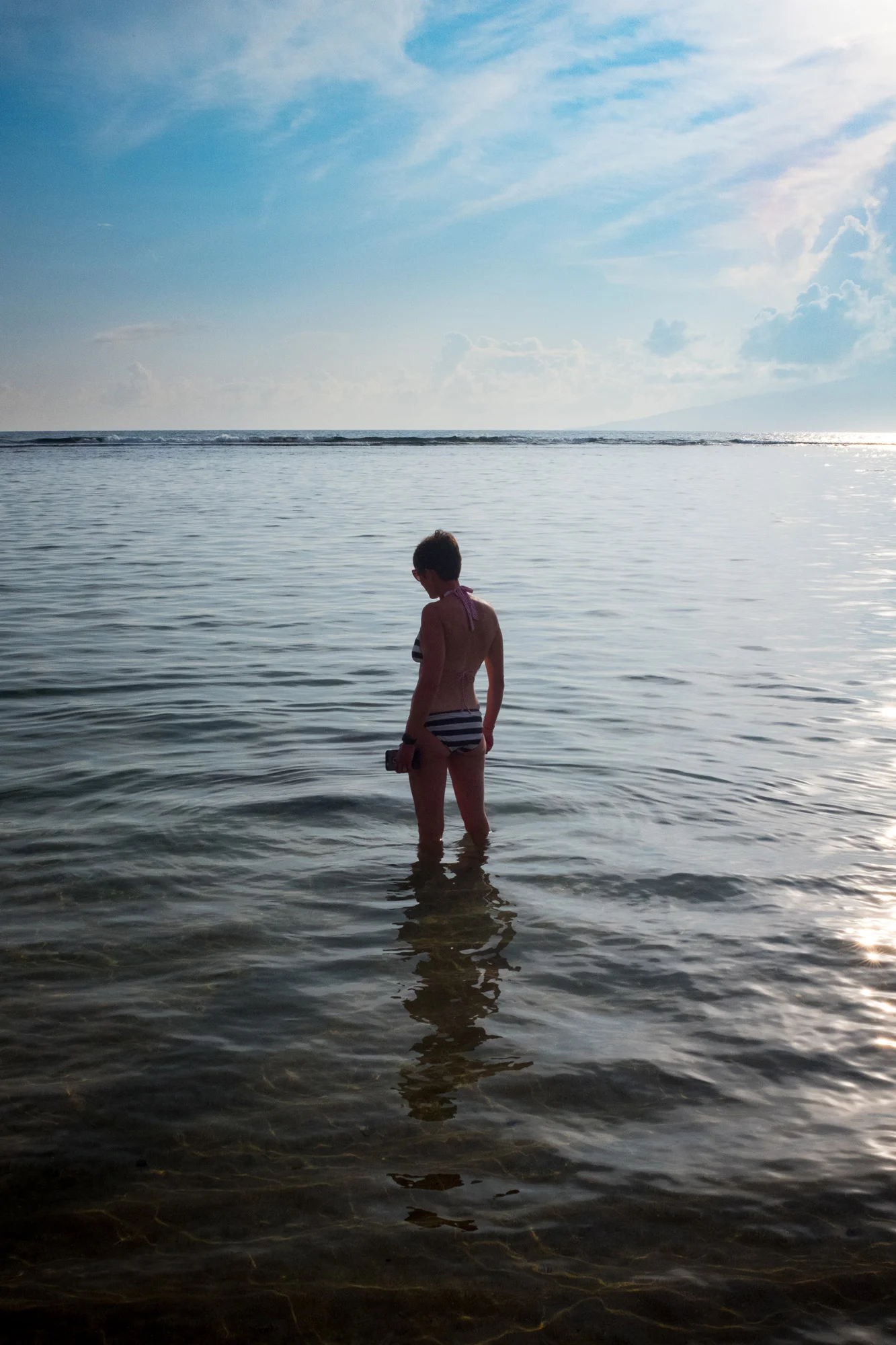 A person standing in shallow water at the beach during sunset, facing away from the camera, wearing striped swimwear and holding an object, with the sky partly cloudy in Maui.