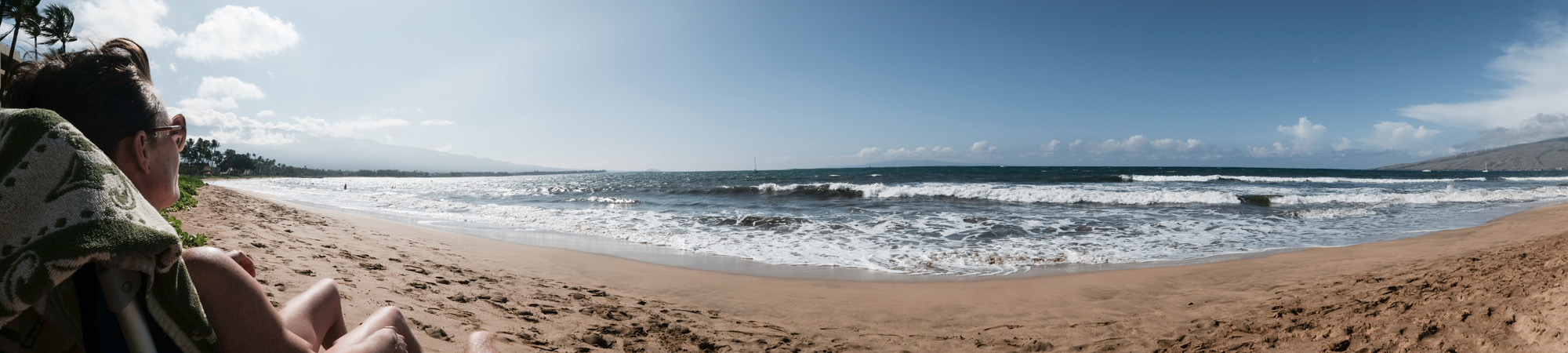 Person sitting on the sandy beach near the shoreline, looking at the ocean with waves and a cloudy sky in Maui.