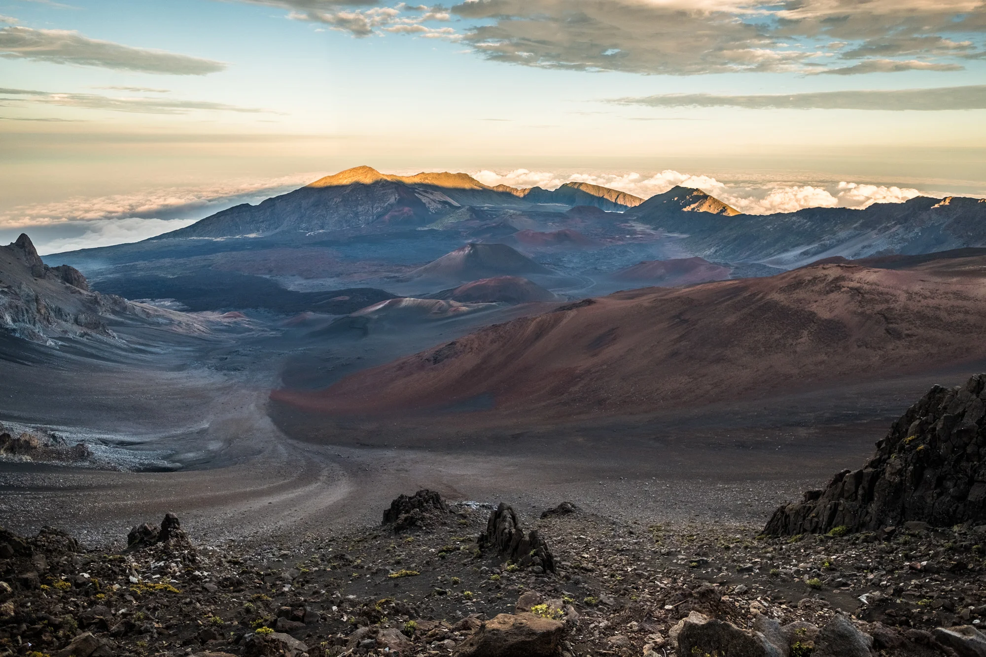A volcanic landscape with rugged terrain, mountains, and clouds in the background, illuminated by soft sunlight Maui.