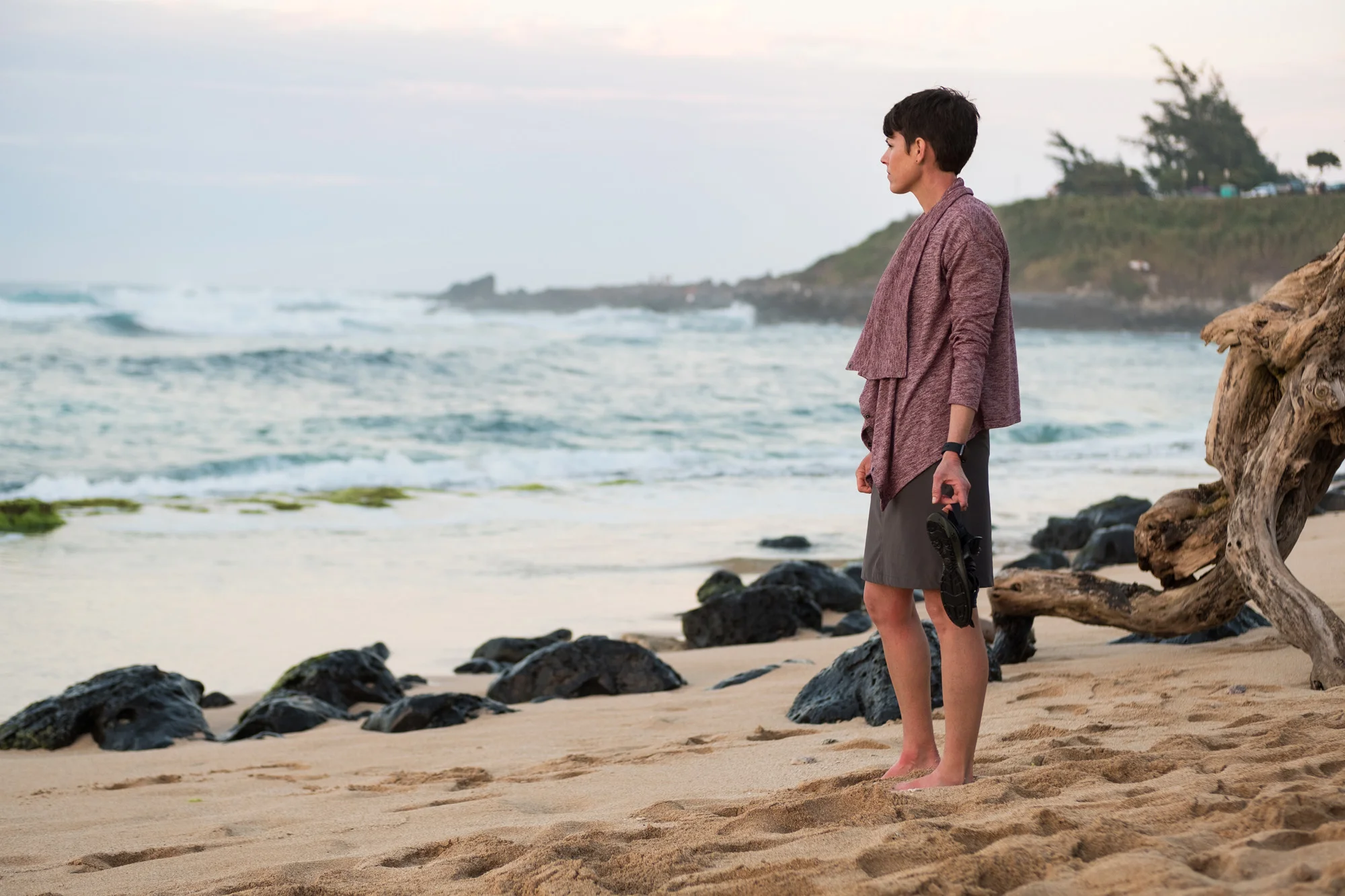 A woman standing on a sandy beach near rocks and driftwood, looking out at the ocean during sunset in Maui.