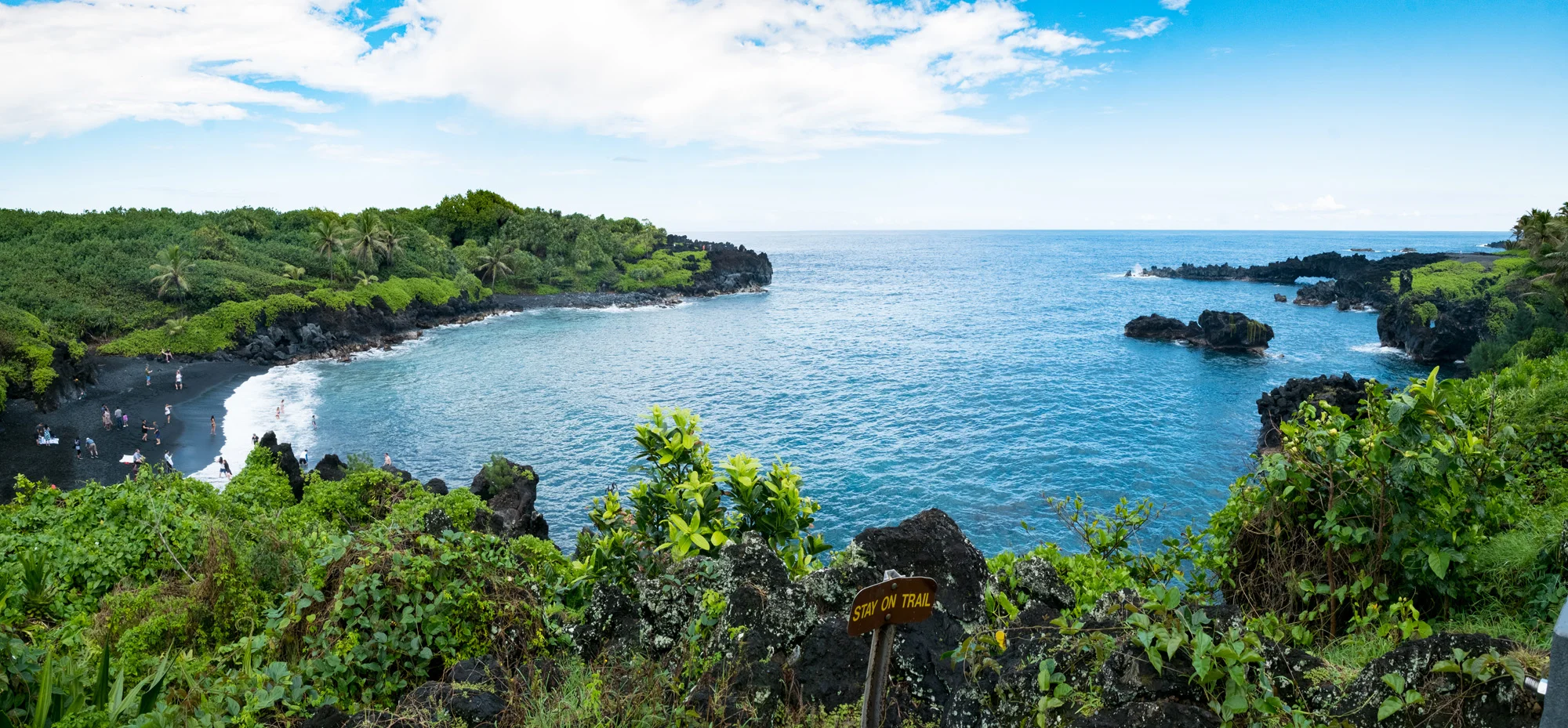 A coastal scene with a black sand beach, lush green vegetation, and rocky cliffs. There are people on the beach and in the water, with a sign that says 'Stay on Trail' in the foreground  in Maui.