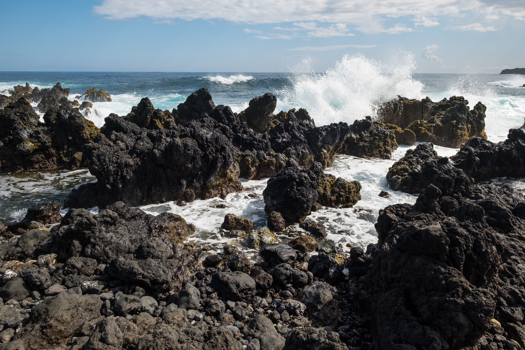 Ocean waves crashing against dark volcanic rocks on a sunny day with blue sky and scattered clouds in Maui.