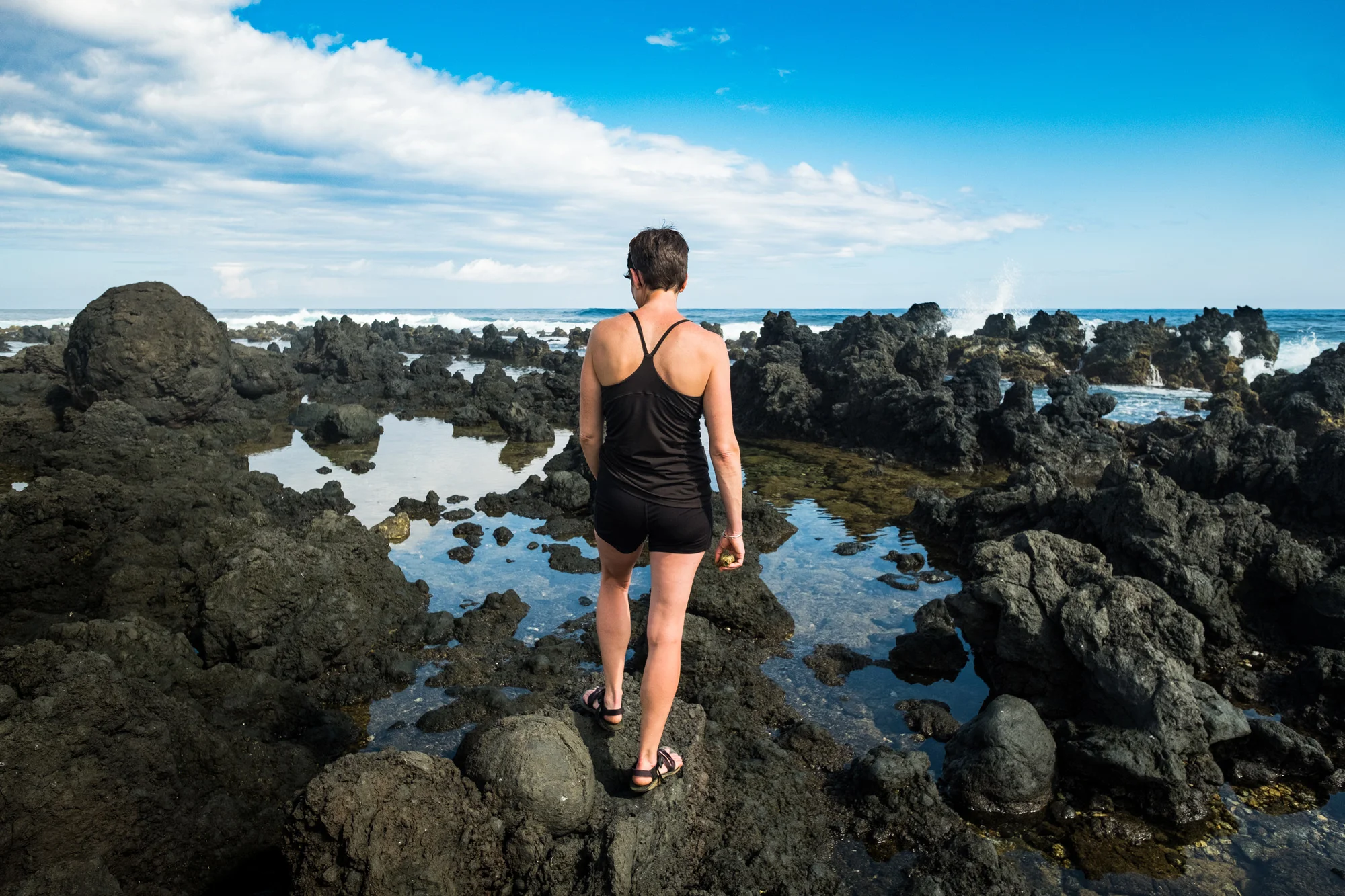 A woman in a black tank top and shorts walking on rocky tide pools at the beach, looking toward the ocean under a partly cloudy sky  in Maui.