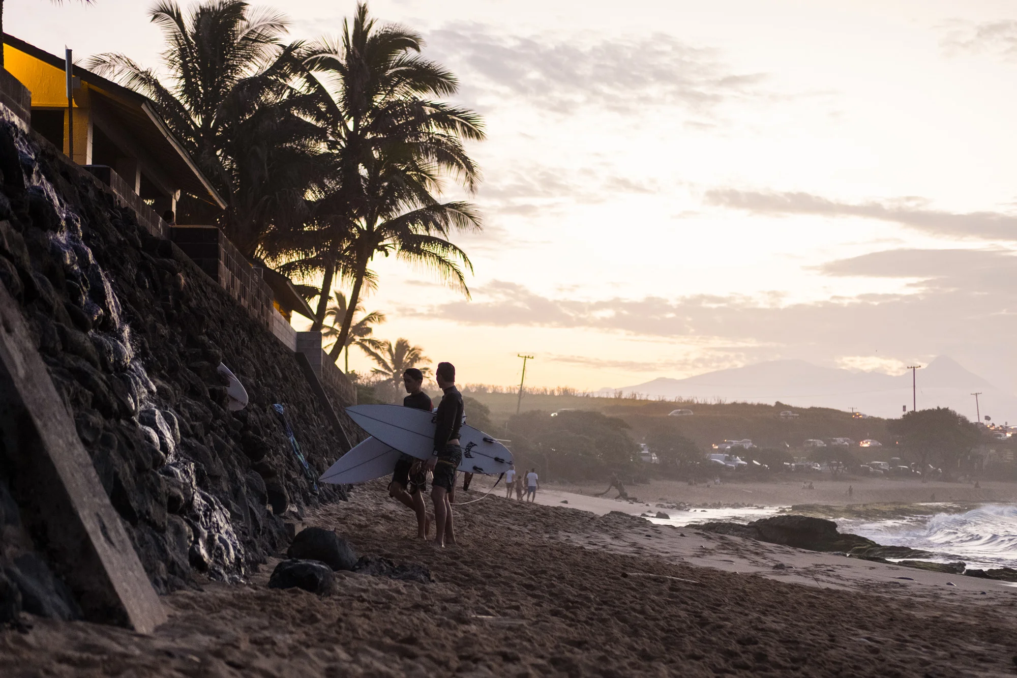 People with surfboards walking along the beach at sunset with palm trees, a stone wall, and distant mountains in the background Maui.