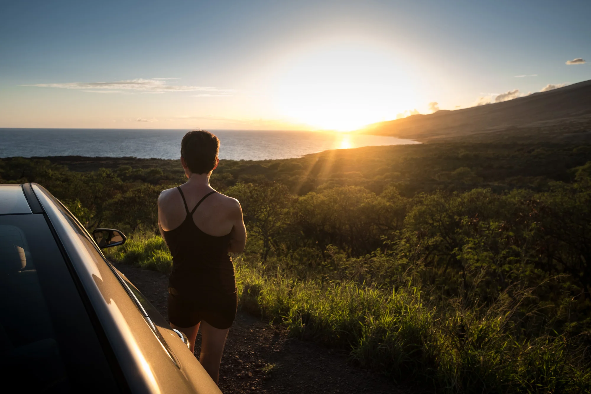A person with short dark hair wearing a black tank top and black shorts standing beside a parked silver car, looking at a sunset over the ocean and hills in the distance  in Maui.