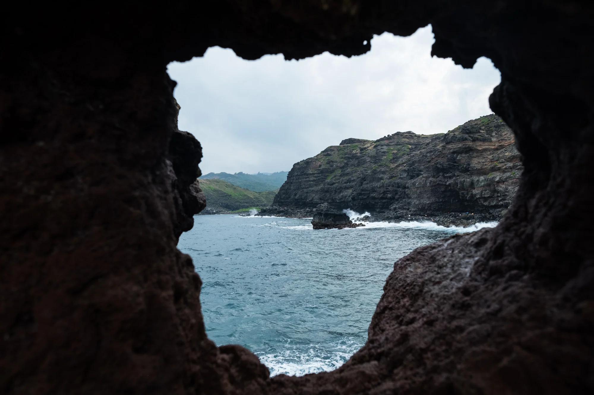 View through a rocky opening of the ocean with waves crashing against cliffs and a cloudy sky overhead Maui.