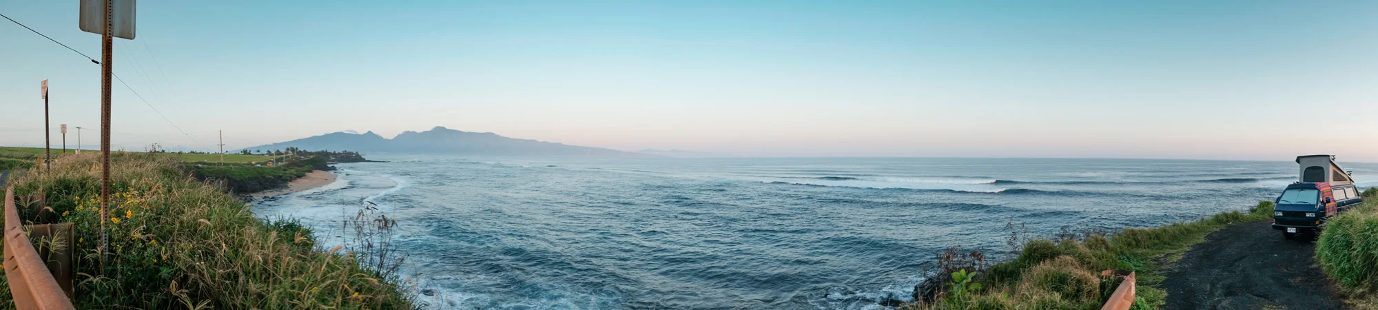 Panoramic view of a coastal landscape with a calm ocean, distant mountains, and a partly cloudy sky. On the right, there is a black camper van parked on a gravel path, with green grass and vegetation nearby. On the left, a small section of a guardrai