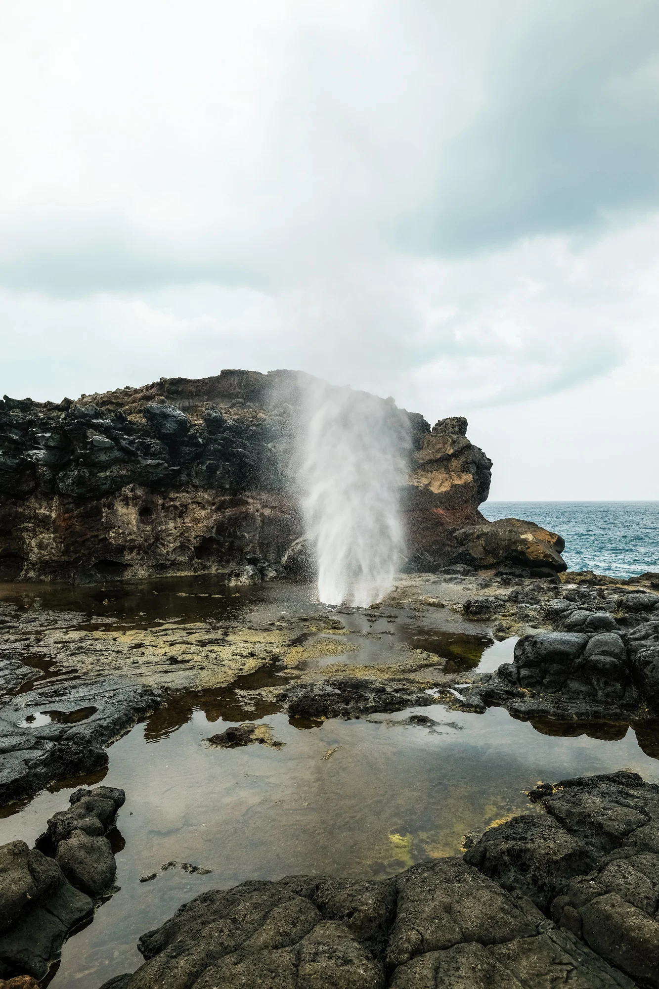 A rocky shoreline with water erupting from a small volcanic crack, with the ocean in the background under an overcast sky Maui.