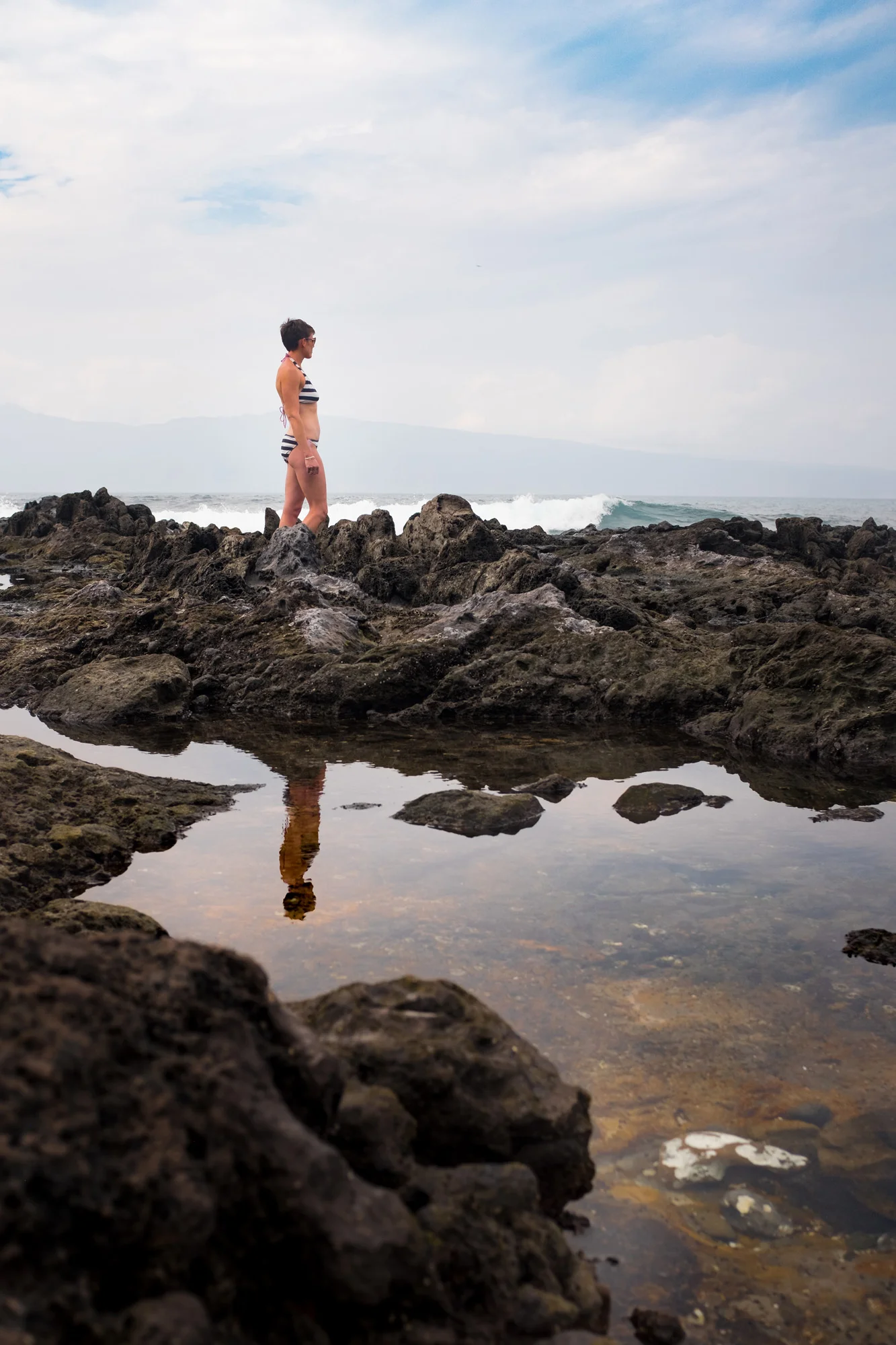 A woman in a striped bikini standing on rocks by the ocean, with her reflection in a small pool of water, under a partly cloudy sky  in Maui.