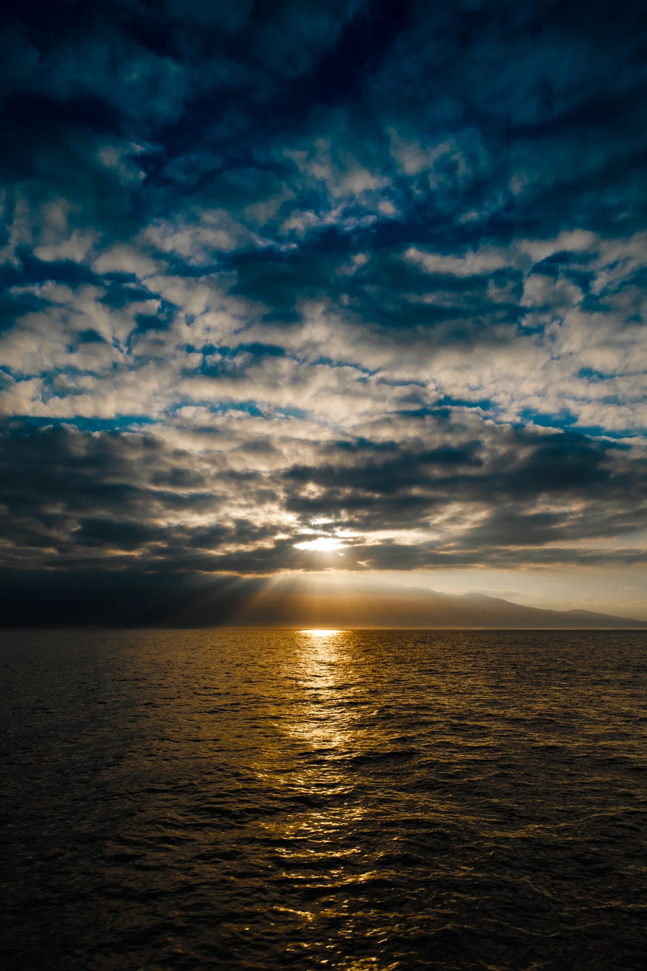 Sunset over the ocean with a dramatic sky filled with dark clouds and rays of sunlight breaking through In Maui.