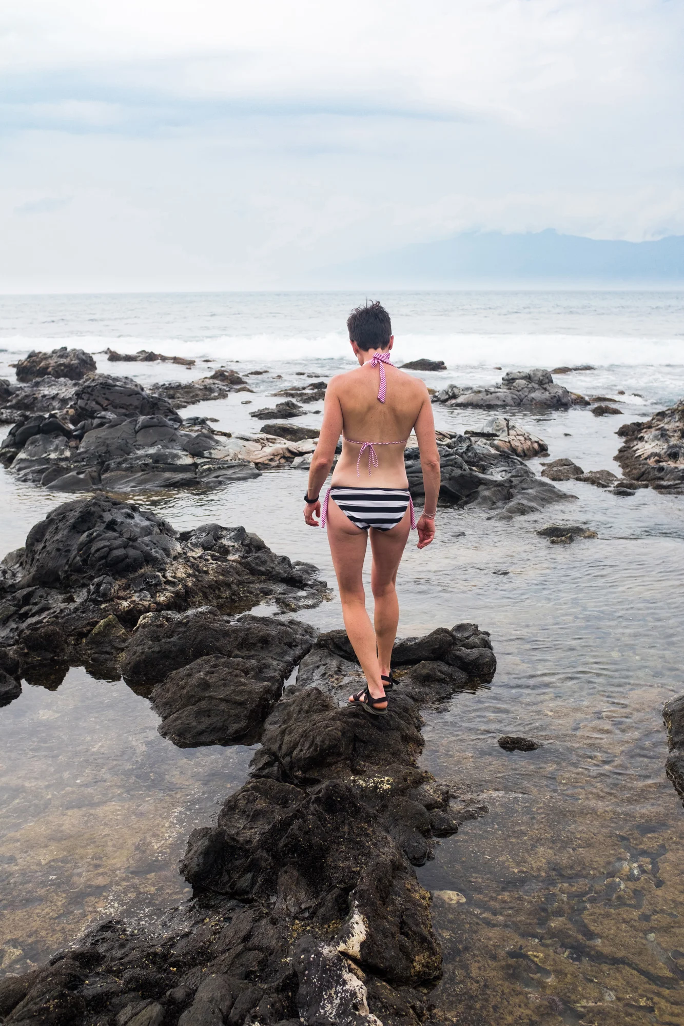 A person with short dark hair wearing a striped swimsuit and black sandals walking on rocks into the ocean, with cloudy sky and distant mountains in the background.
