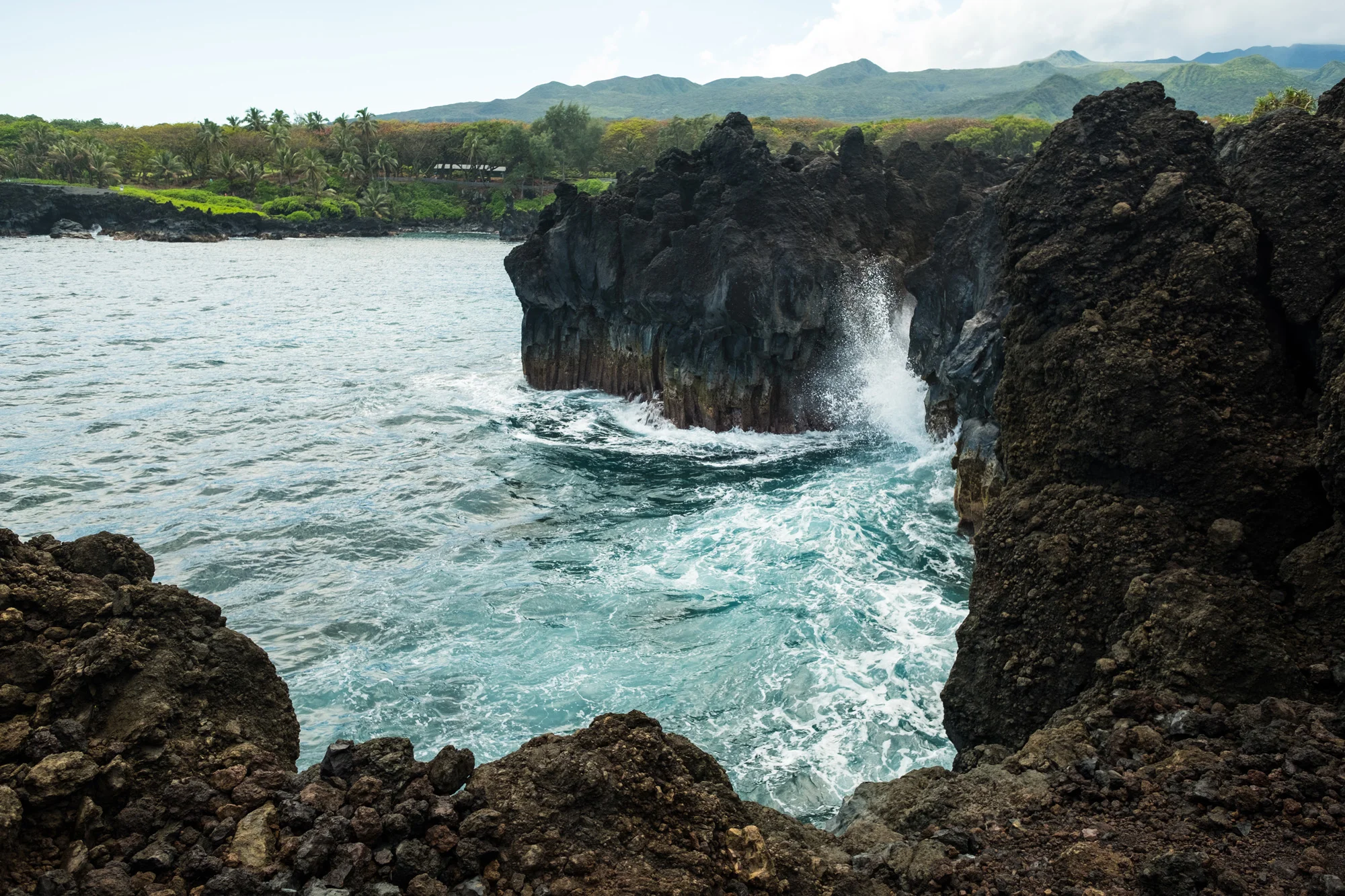 Black volcanic rocks along the shoreline with waves crashing against them, lush green trees, and mountains in the background under a cloudy sky  in Maui.